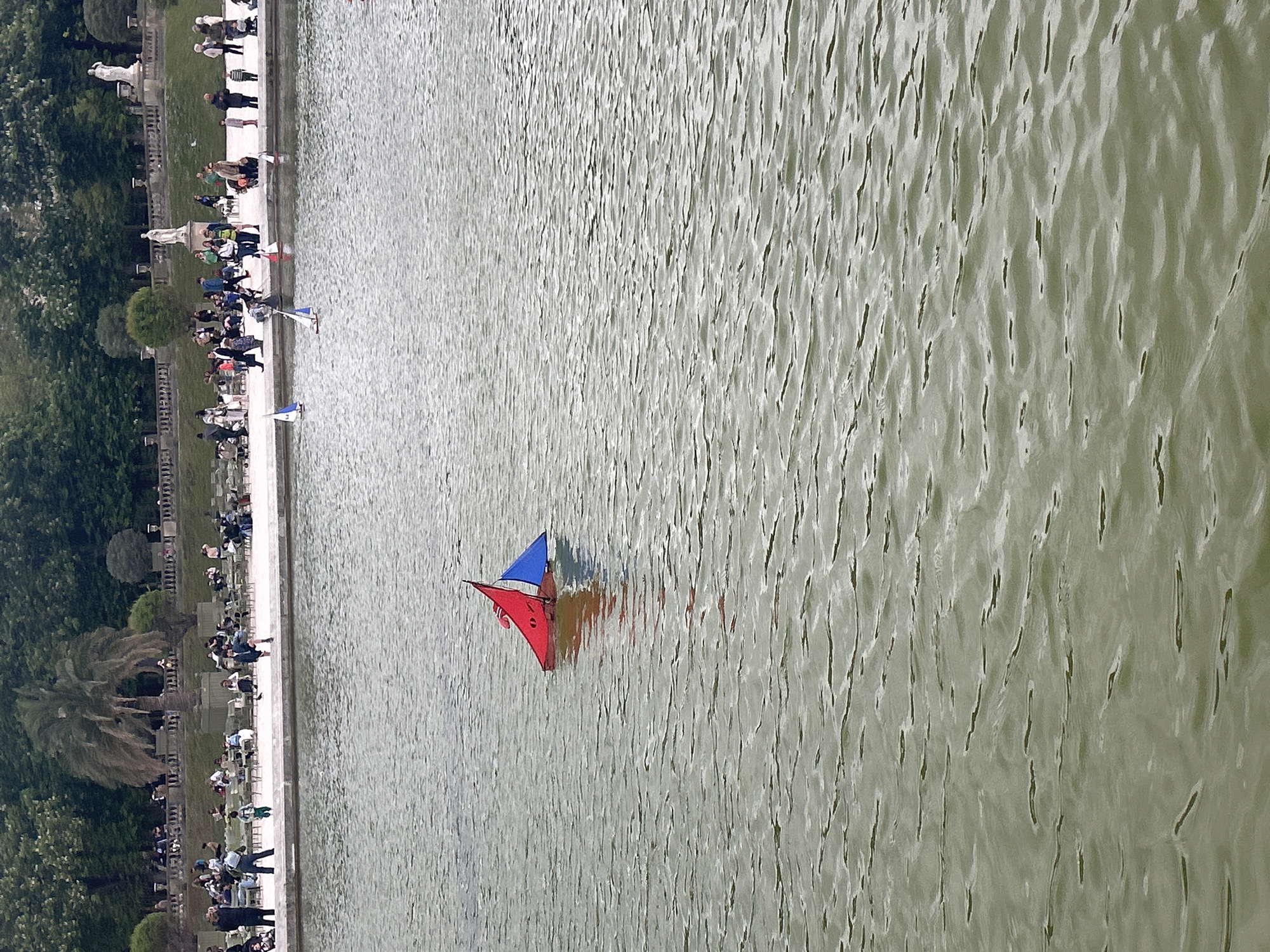 The image depicts the Luxembourg Garden in Paris, France. The focal point is a pond with a toy sailboat featuring red and blue sails. The water surface is rippled. Along the edge of the pond, a line of people sits and stands, watching the boats. The shoreline is lined with trees and garden vegetation. A stone balustrade separates the walkway and the garden. Statues and trees are interspersed along the pond's edge. The image suggests a calm, leisurely atmosphere.