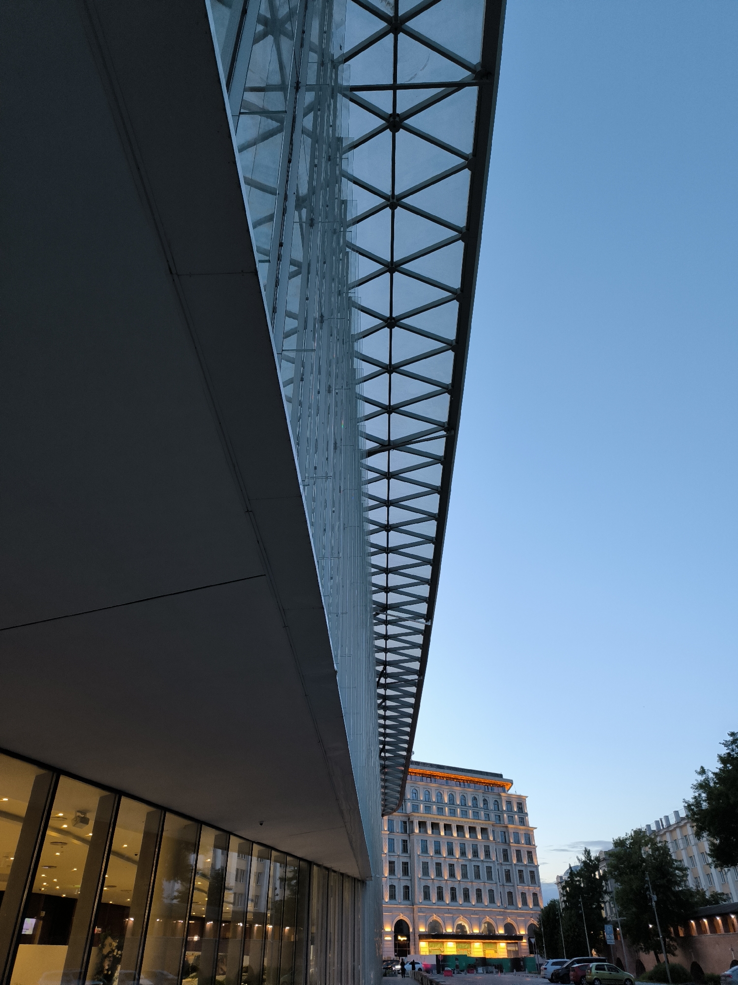 An exterior shot of a modern building with a triangular glass roof structure extends into a clear blue sky. The building's facade features large, reflective glass windows, revealing interior lighting and architectural elements. To the right, a multi-story building with a light-colored facade and numerous windows is visible, illuminated by interior and exterior lights. Trees line the street in front of this building. A few vehicles and pedestrians are present on the street. The overall impression is an urban environment at dusk or early evening, likely in Moscow, Russia, given the architectural style.
