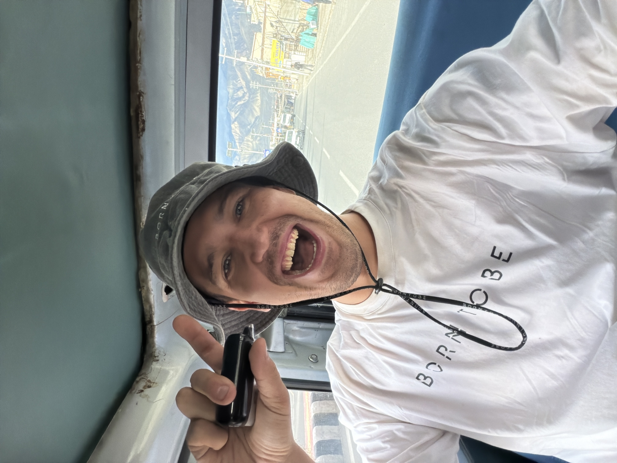A man wearing a grey bucket hat and a white t-shirt with "BORN TO BE" printed on it is taking a selfie inside a vehicle, possibly a bus or van. He is smiling and holding up a black object, possibly an electronic device. The background shows a street scene in Leh, India, with buildings, vehicles, and mountains in the distance. The interior of the vehicle appears somewhat worn, with stained headliner. The man's shirt has a drawstring attached to the hat.