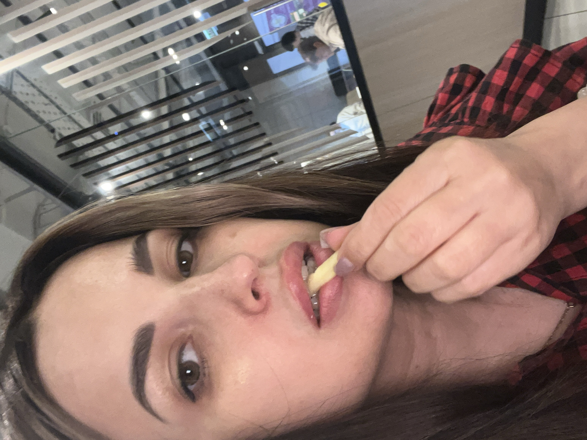 A woman with brown hair and fair skin is holding a french fry to her mouth. She is wearing a red and black plaid shirt. The background shows a ceiling with a linear pattern and a person in a blurred reflection. The location is Unknown, Unknown.