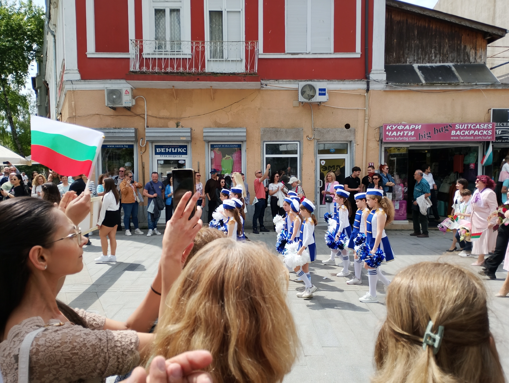 A street scene in Burgas, Bulgaria, shows a parade or performance involving young girls dressed in blue and white sailor-style outfits and white pom-poms. They stand in formation, likely performing a routine, as spectators watch and take photos. A woman on the left holds a Bulgarian flag. The crowd includes people of various ages. Buildings line the street, with shops visible in the background. The shops have signs in Bulgarian, including "ВЕНИКС ПРОИЗВОДИТЕЛ" and a sign for "КУФАРИ ЧАНТИ big bags SUITCASES BACKPACKS". An air conditioning unit is visible on one of the buildings. The street is paved with grey stone. The overall setting suggests a public celebration or local event.