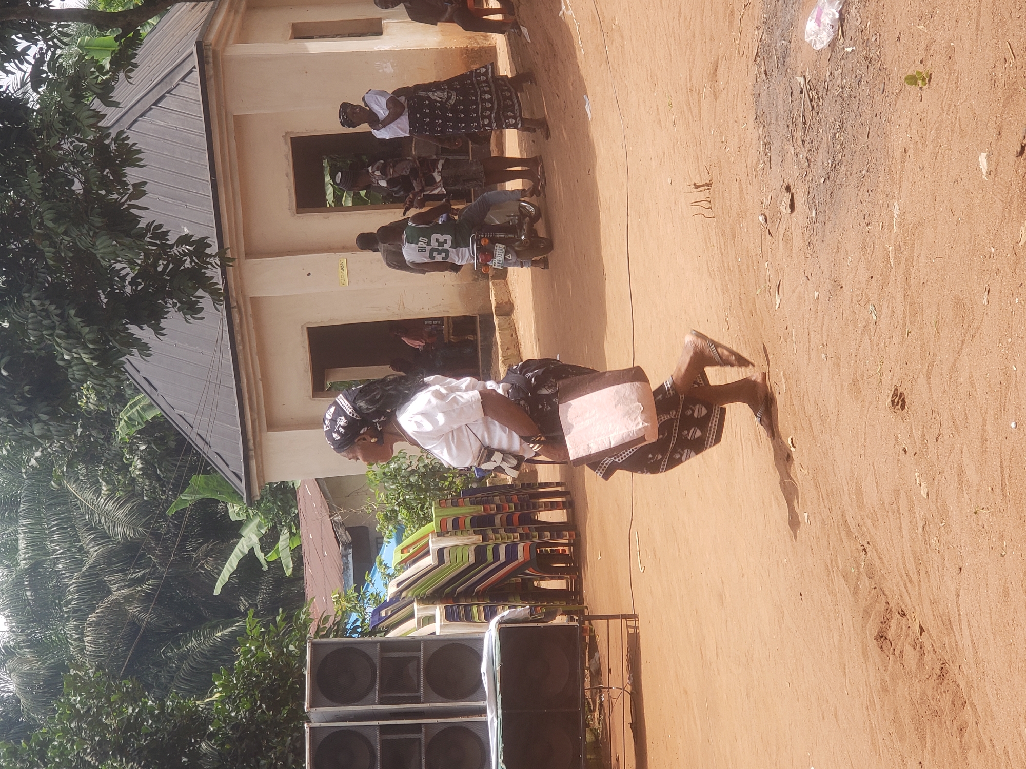 The image depicts an outdoor scene in Umundugba, Nigeria. A woman is walking on a sandy surface. She is wearing a white top, a dark patterned skirt, a head wrap, and sandals, and carrying a pink bag. A stack of colorful plastic chairs is visible behind her, along with several large speakers. In the background, there is a building with beige walls, some individuals standing in front of it, and a motorcycle parked nearby. A tree with lush green foliage is on the left side of the frame.