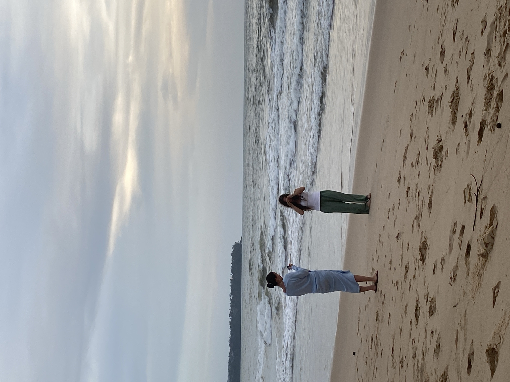 Two women are standing on a sandy beach in Choeng Thale, Thailand, near the water's edge. One woman is wearing a light blue outfit, and the other is wearing a white top and green pants. The sky is cloudy, and the waves are visible in the background. Footprints cover the sand on the beach.