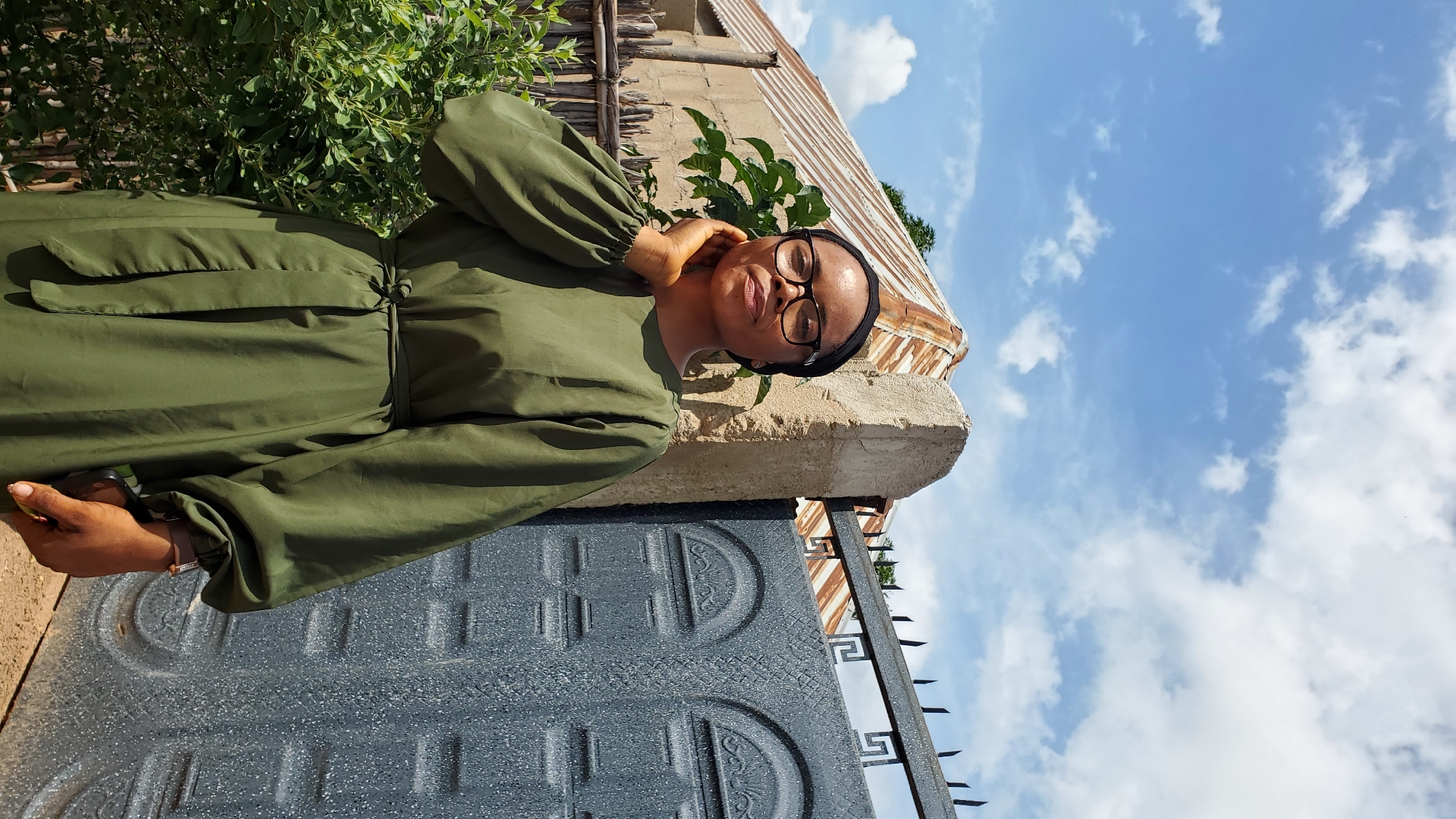 A young woman wearing a green dress and a black headscarf stands outdoors in Jalingo, Nigeria. She is looking forward with a slight smile and her left hand is touching her neck. Behind her is a building with a corrugated metal roof and a gray metal gate. There are trees and bushes to the left of the woman. The sky is blue with white clouds.