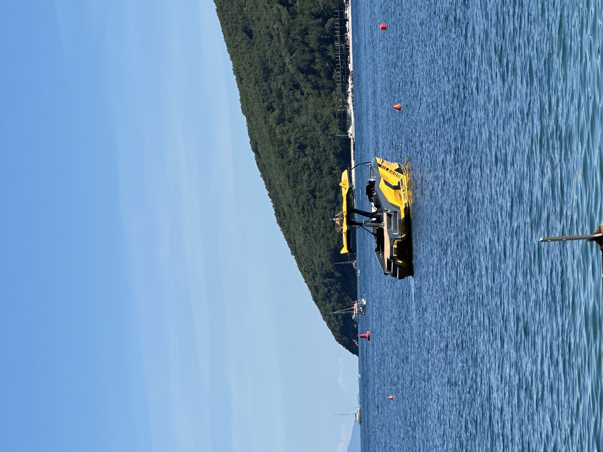 The image shows a yellow watercraft on the water in what is likely the coastal city of Budva, Montenegro, given the presence of the coastline, the green, mountainous landscape in the background, and typical coastal waters. The watercraft is in the center of the frame. There are several red navigation buoys in the water. The sky is clear and blue, indicating a sunny day. There is a wooden post/beam visible in the lower right corner.