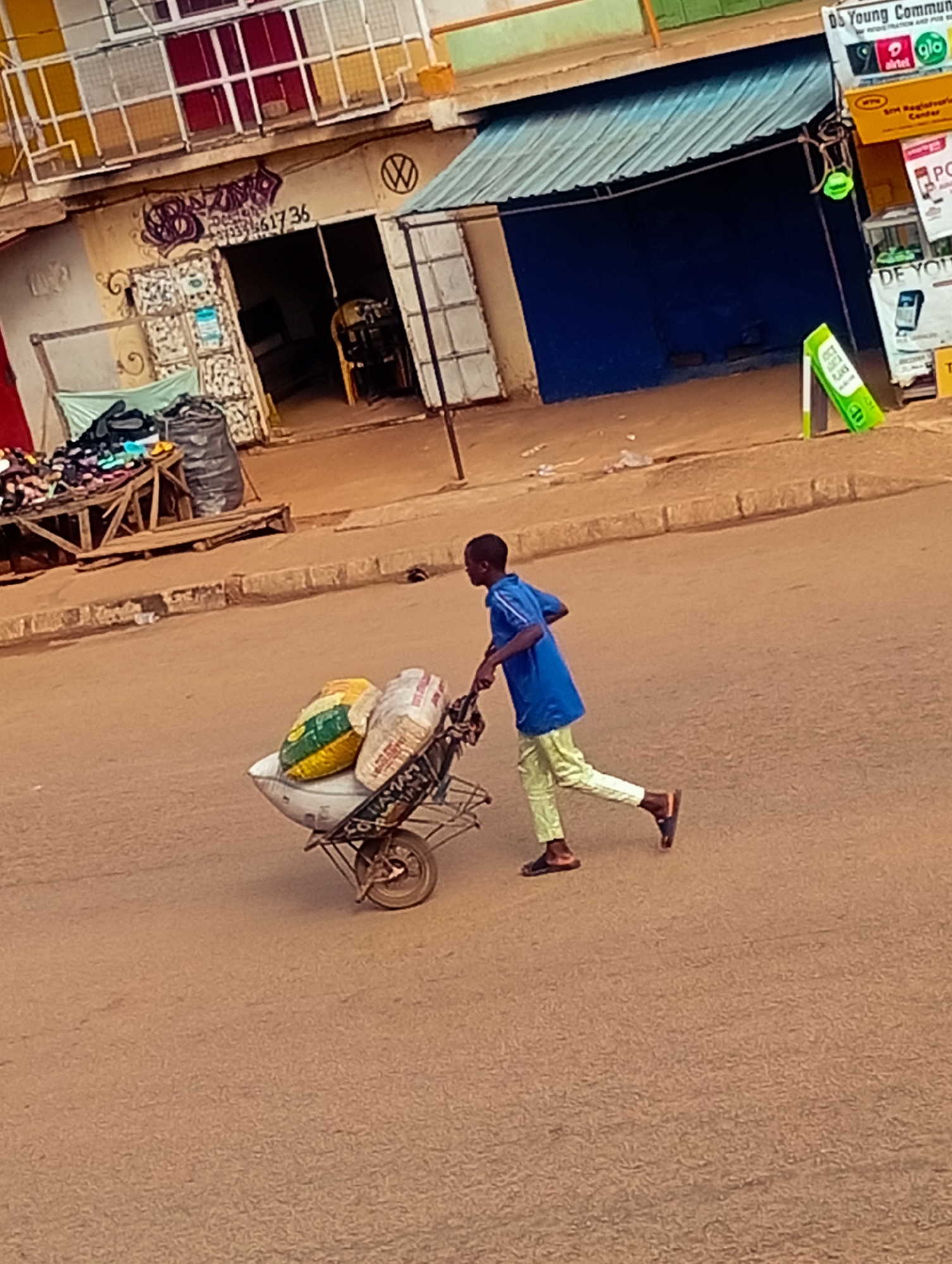 In Bukuru, Nigeria, a young boy, wearing a blue t-shirt and light green pants, is pushing a wheelbarrow loaded with three large sacks of grain across a paved street. The wheelbarrow has a single wheel in the front. The boy is moving from left to right in the frame. Behind him, a row of shops with open entrances and closed shutters line the street. One shop features a prominent yellow sign with telecommunication provider logos, while another has graffiti art on its facade. In the foreground, the street is rough and dusty. The scene suggests a typical street activity in a Nigerian city.