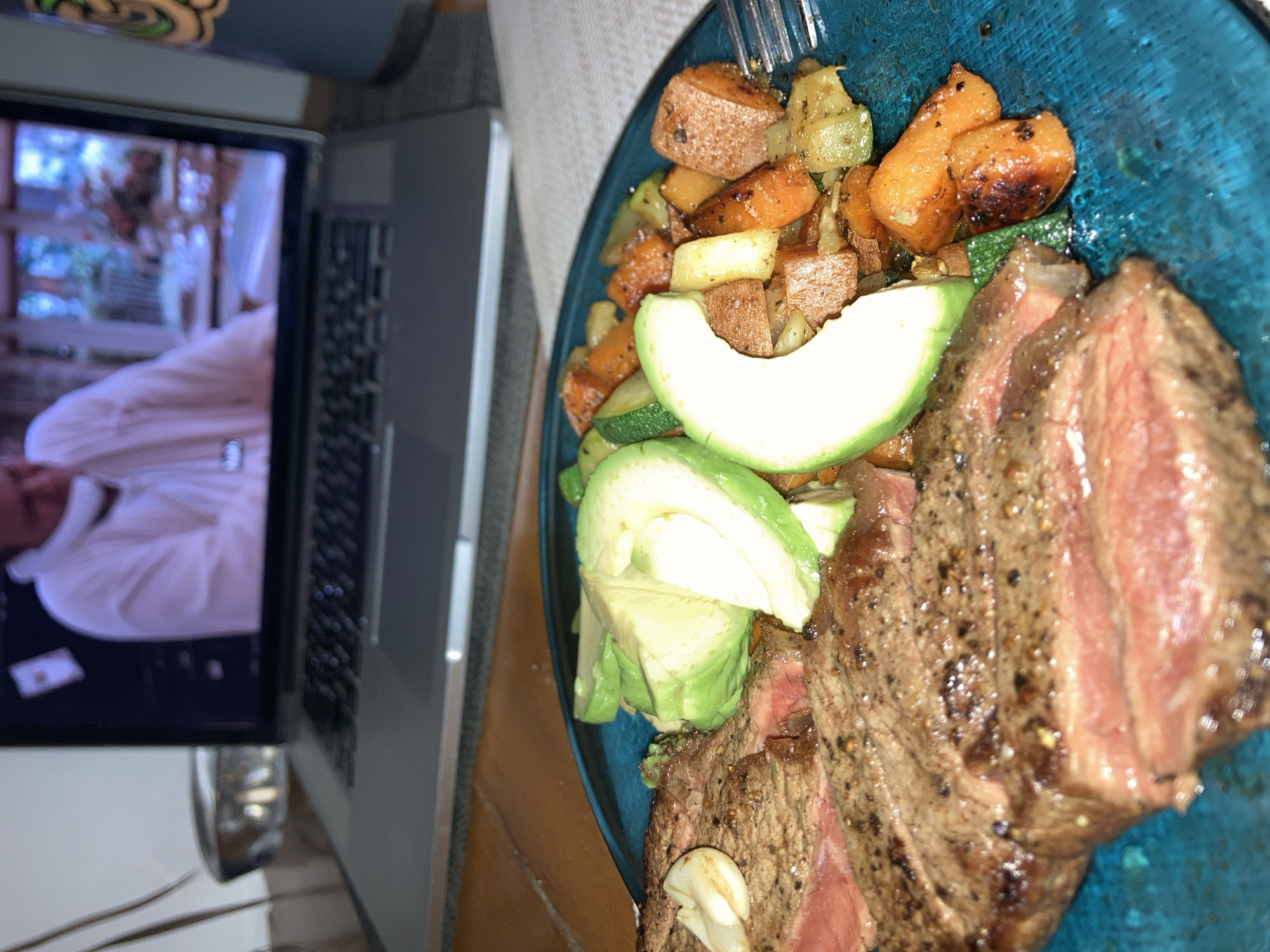 A plate of food is placed next to a laptop. The food consists of cooked steak, avocado slices, and roasted vegetables. The laptop screen displays a video. The setting appears to be indoors. There is no indication of the image's location in Barcelona, Spain.