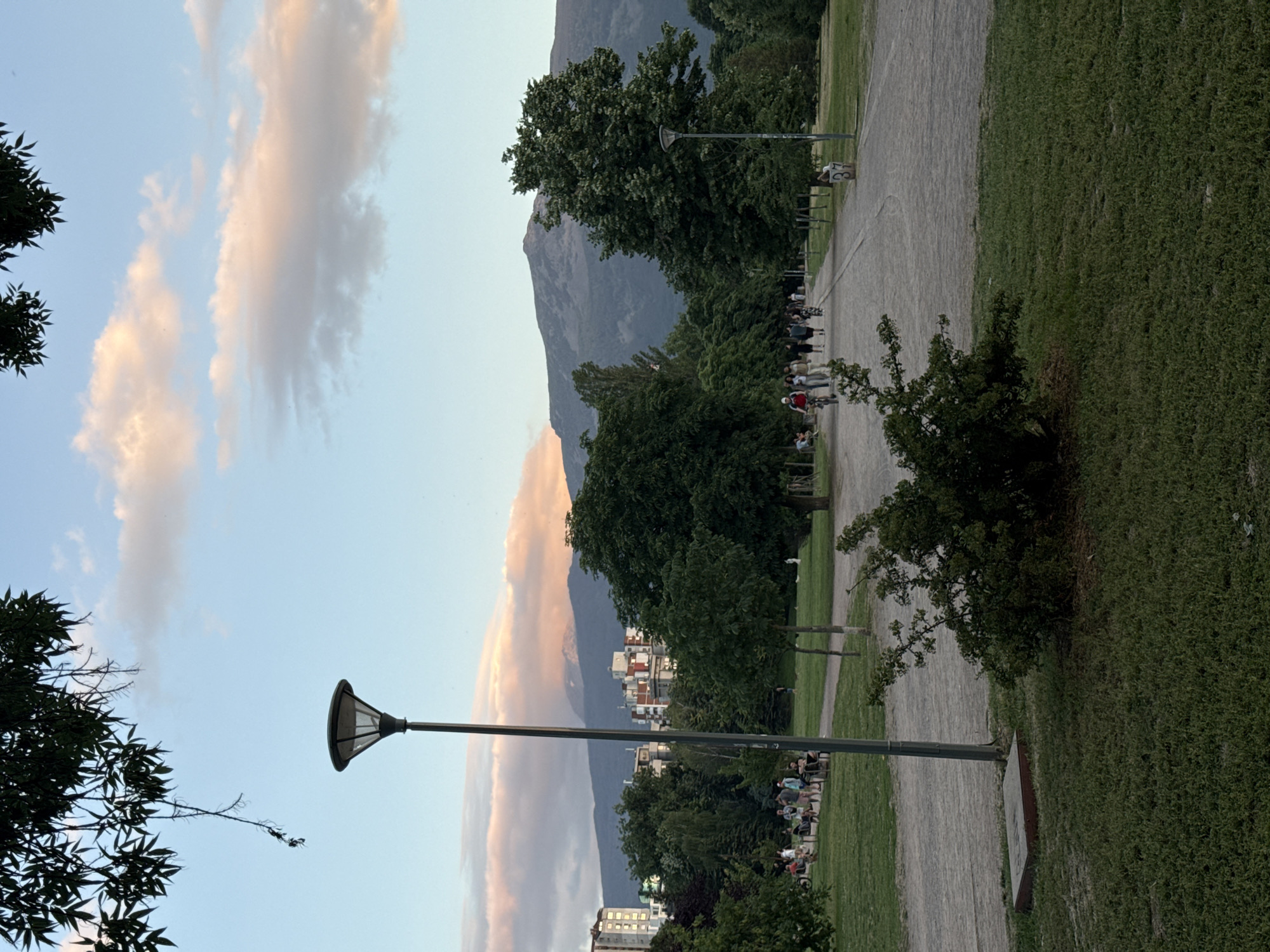 This image depicts a view of Vitosha Mountain from the Borisova Gradina park in Sofia, Bulgaria. The foreground is dominated by a well-maintained lawn and a gravel pathway. A contemporary-style lamppost stands on the left side of the path. In the mid-ground, several people are walking and cycling along the pathway, with some sitting on benches. Dense green trees line both sides of the path, with apartment buildings visible in the background beyond the trees on the left. The background features the imposing silhouette of Vitosha Mountain, with a soft, pinkish glow from the setting sun illuminating some of the clouds above it. The sky transitions from a pale blue at the top to a warmer hue near the horizon. The overall atmosphere is peaceful and serene, characteristic of a park at dusk.