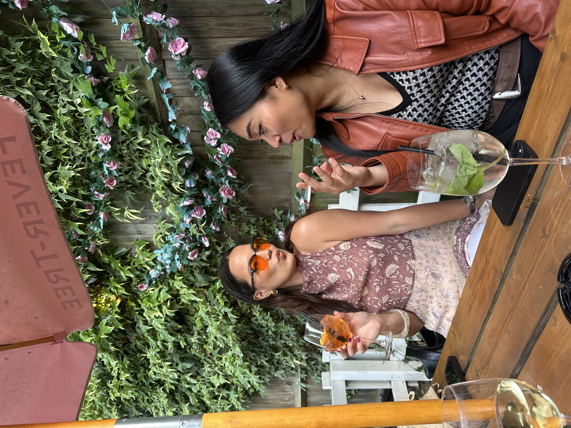 Two women are seated at a wooden table in an outdoor setting. One woman wears a brown leather jacket and a black and white patterned top, while the other wears a printed dress and orange sunglasses. Both are holding glasses of drinks. The surrounding environment includes lush greenery, with flowers and vines climbing a wooden fence. The scene is illuminated by natural light. A sign appears in the bottom left corner of the image that spells out "LEAVE". The location is likely a restaurant or bar in the city of London, United Kingdom.