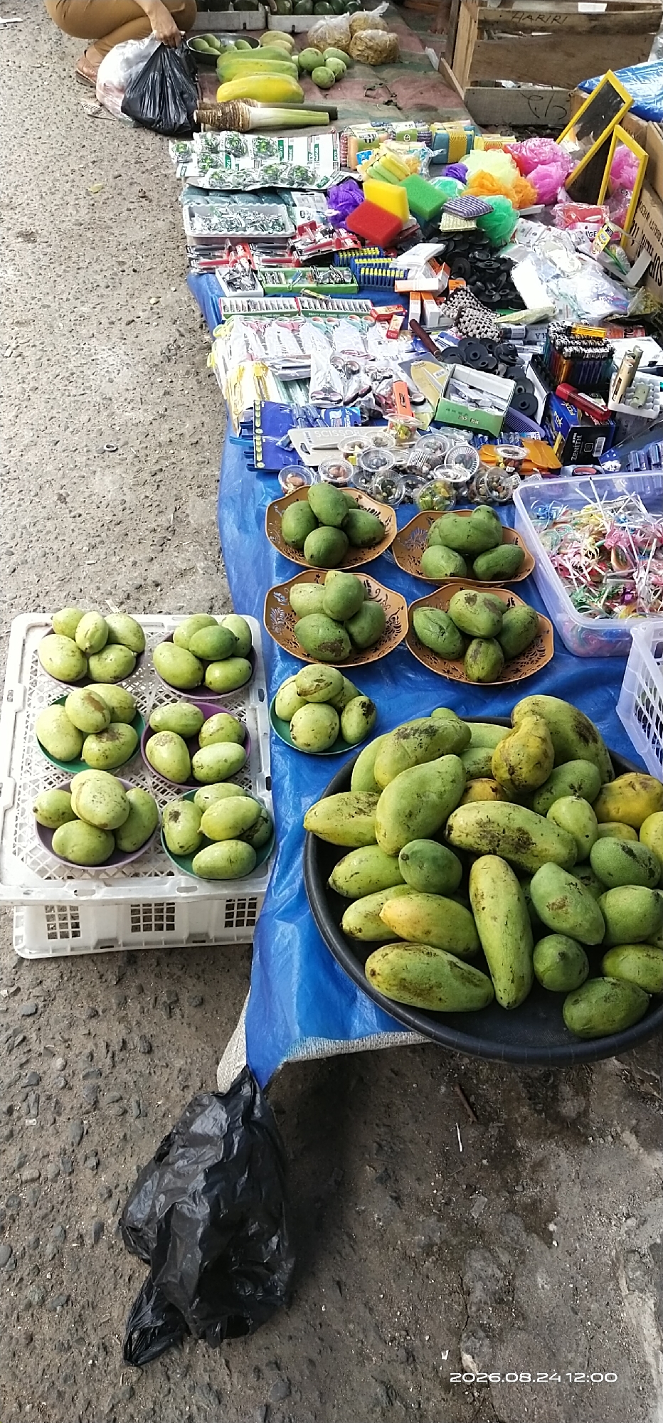An overhead shot captures a marketplace stall in Tamiang Layang, Indonesia, displaying a variety of fruits and small goods. In the foreground, a black plastic bag lies on the concrete ground. To the left, a white plastic crate contains several piles of green mangoes. Adjacent to it, on a blue tarp, are multiple brown bowls filled with more green mangoes, with a large black circular tray overflowing with mangoes to the right. The stall itself is laden with colorful sponges, packaged soaps, and other small items. The ground is composed of coarse gravel and dirt.