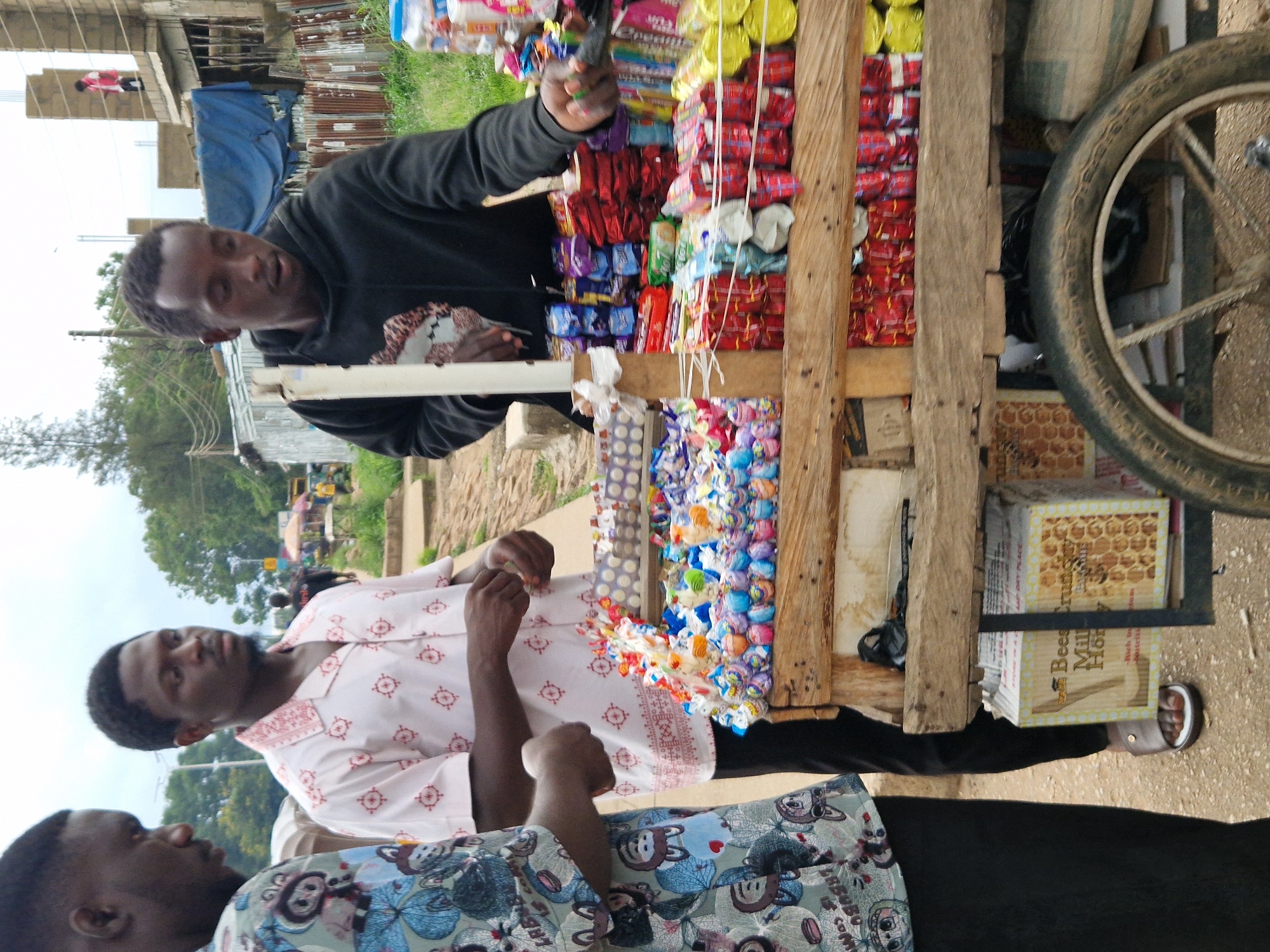 The image depicts a street scene in Jos, Nigeria, focused on a man operating a candy cart. The cart, made of rustic wood, is laden with a variety of colorful candies and packaged goods. Two other men are present, one standing to the left and another in the background, partially obscured. The man at the cart, wearing a black hoodie, appears to be interacting with customers or arranging his merchandise. The background reveals a bustling urban environment with buildings under construction, corrugated iron fencing, and a cloudy sky. The presence of the cart and its wares suggests a commercial activity taking place in a public space within the city.