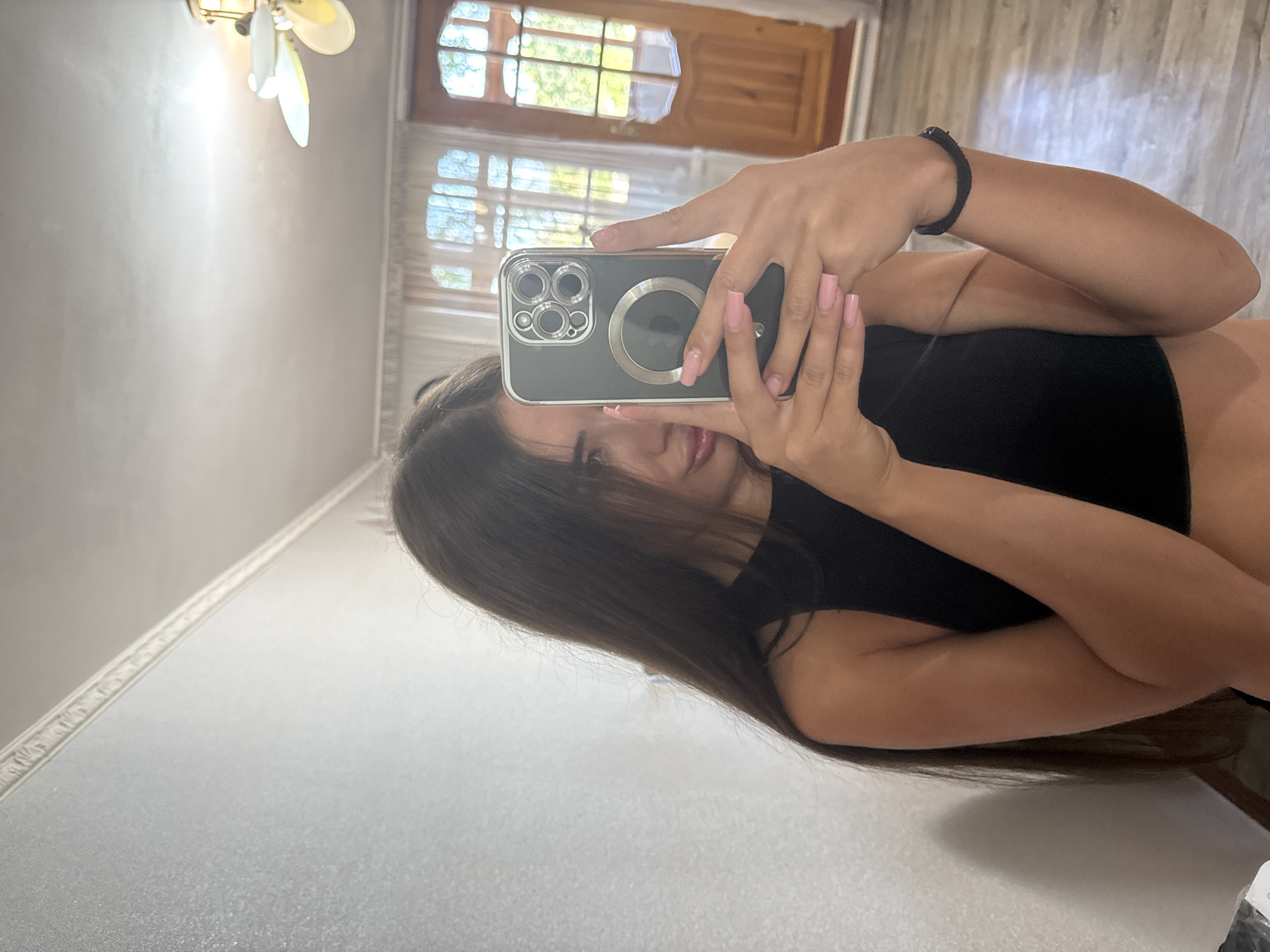 A young woman with long, dark hair is taking a mirror selfie indoors. She is wearing a black, sleeveless top and has long, pink fingernails. Her face is partially obscured by her hair and the phone she is holding. The phone is a dark color with a prominent camera module. The background appears to be a room with light-colored walls, a white ceiling with a decorative molding, and a ceiling fan. A wooden door with windows and sheer curtains are visible in the background. The floor is a light-colored wood or laminate. The overall lighting suggests daytime. There is no identifiable information in the image to suggest the location is Basarabeasca, Moldova.