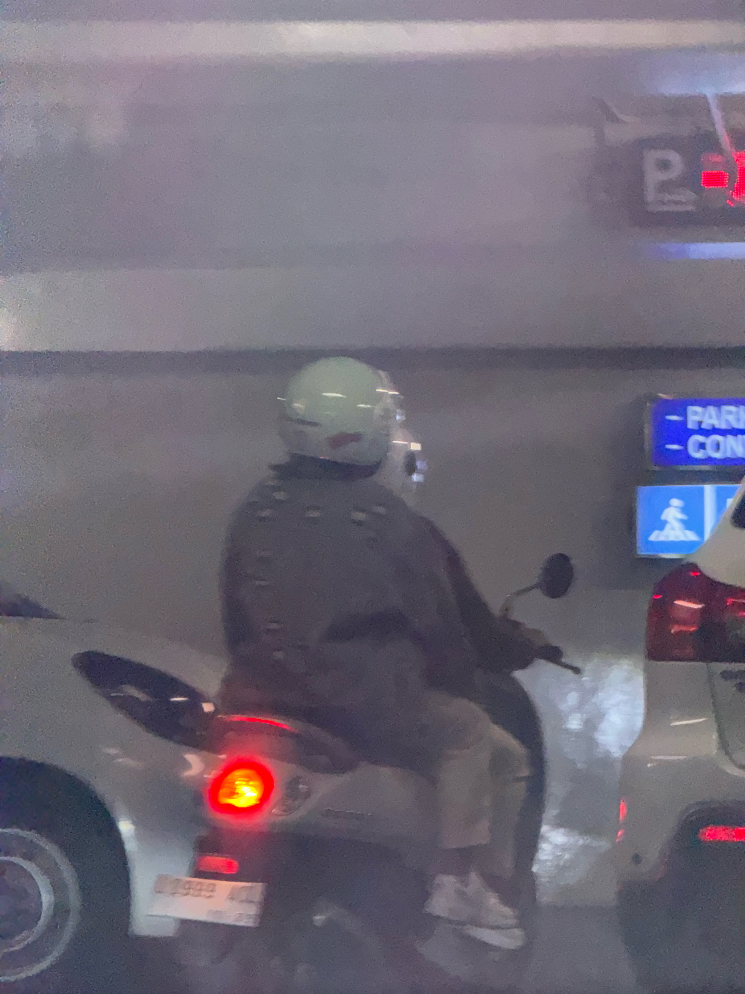 The image captures a person riding a scooter in what appears to be an indoor parking structure. The rider is wearing a light green helmet and a dark, patterned jacket. They are also wearing light-colored pants and white sneakers. The scooter has a red taillight illuminated. In the background, there are signs indicating parking. One sign, in blue, appears to have text related to parking and possibly a contact number. Another blue sign below it displays a pictogram of a person walking. The overall lighting suggests an indoor environment, possibly a dimly lit parking garage. The presence of license plates on vehicles and typical parking signage suggests an urban setting, and given the provided context, likely Surabaya, Indonesia.