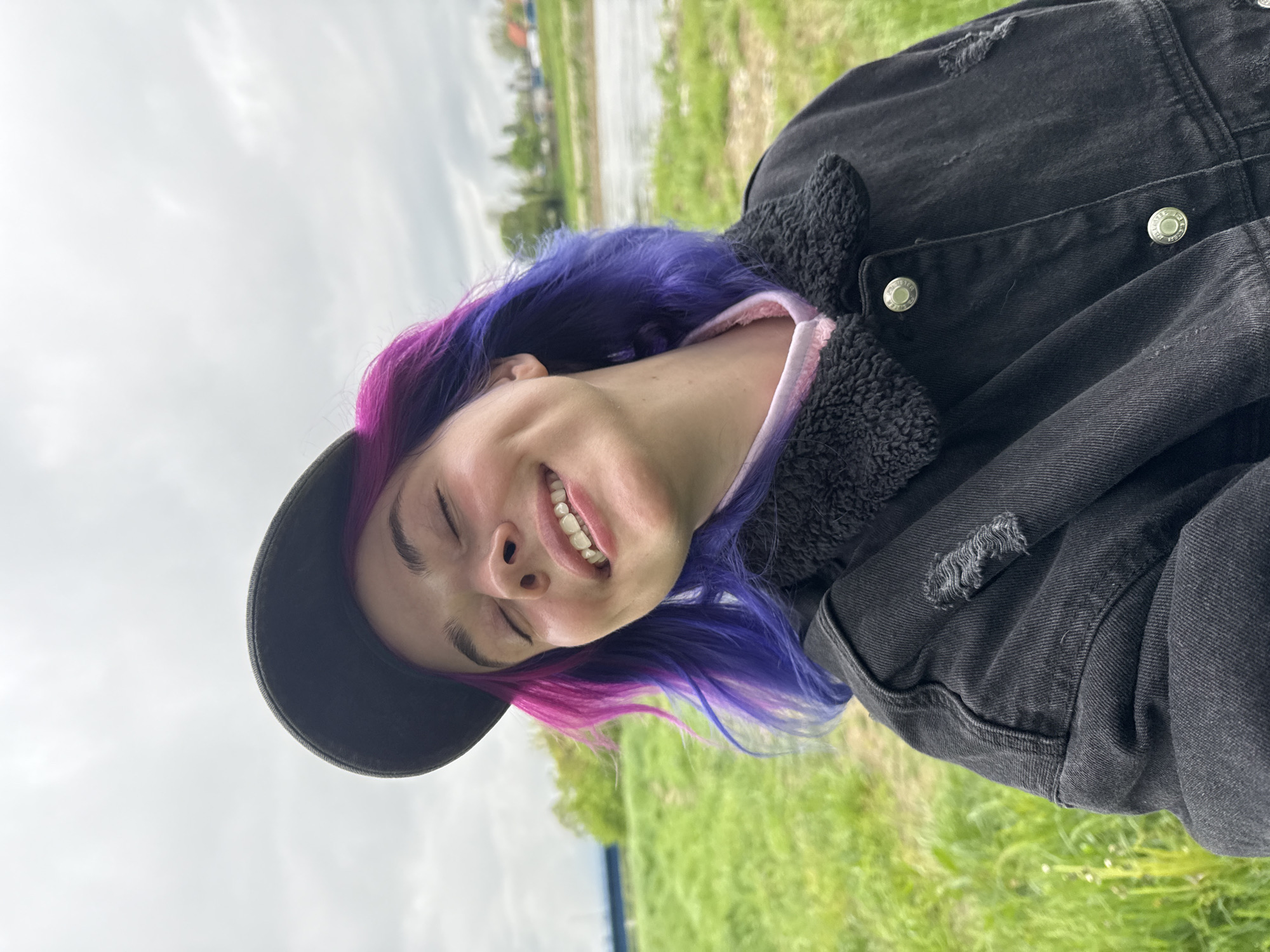 A person with purple and pink hair and a black baseball cap smiles with their eyes closed. They are wearing a black denim jacket with a sherpa collar and a pink top underneath. The background consists of green grass and a cloudy sky. The location is outside and possibly near a body of water. The image appears to be taken in Croatia, likely in the City of Zagreb.