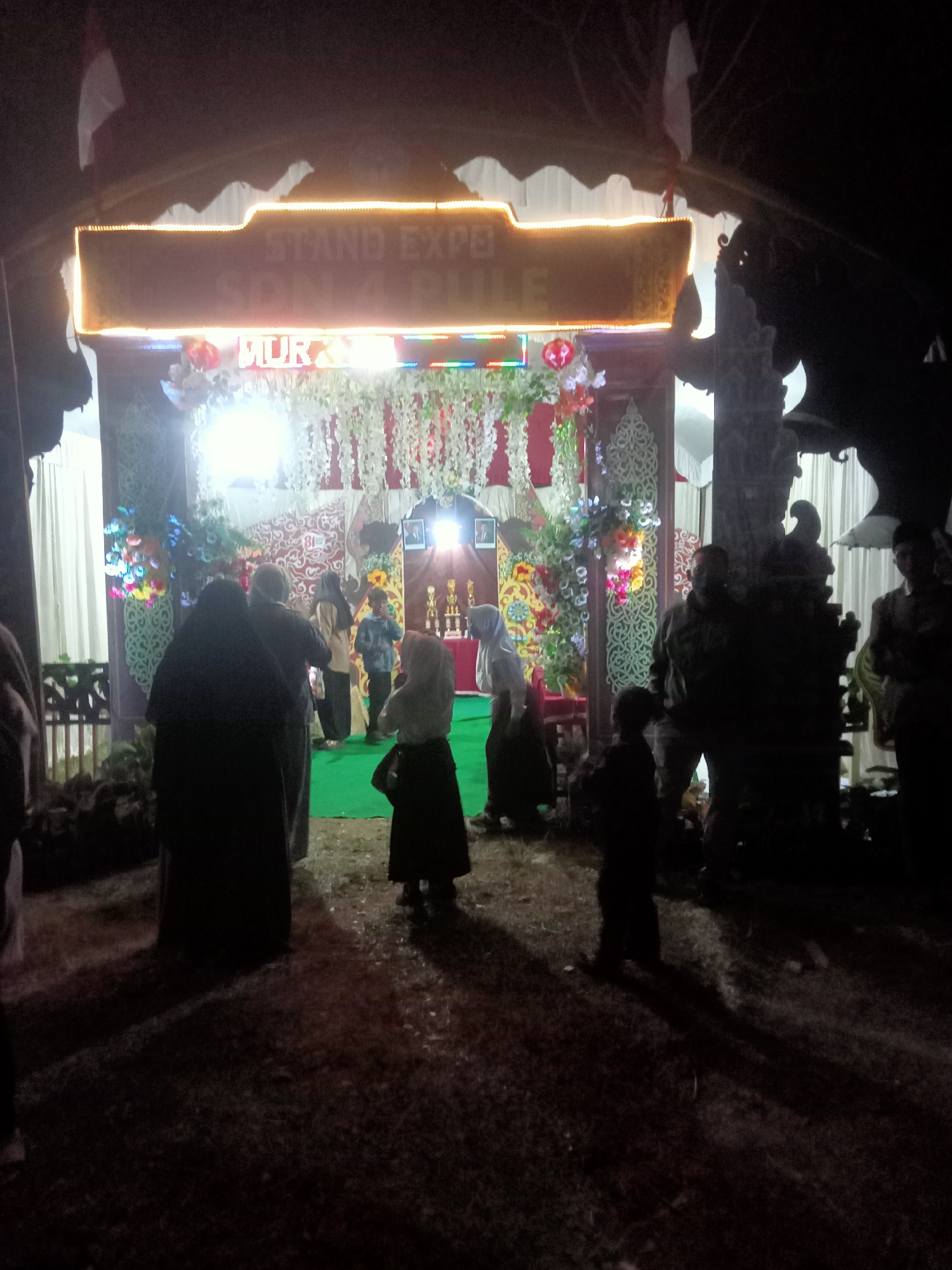 The image captures an outdoor event at night, possibly a stand expo or similar gathering, in the city of Pule, Indonesia. A brightly lit archway, adorned with what appear to be white artificial flowers and colorful lights, marks the entrance to a decorated stall or stage. Text on a banner above the entrance reads "STAND EXPO" and "SDN 4 PULE". Several people, including adults and children, are visible in the foreground and background, some appearing to observe the displays or participate in the event. The ground is covered in grass and dirt. The overall atmosphere suggests a community event taking place in a semi-rural setting.