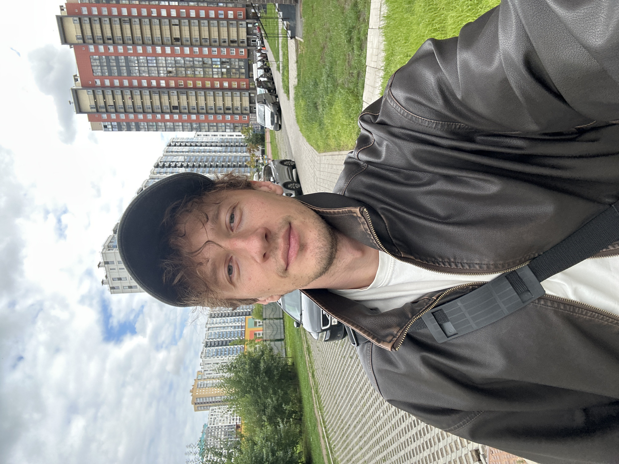 A medium shot captures a man in a dark brown leather jacket and a baseball cap, facing the camera. He has curly brown hair and is looking directly at the lens. Behind him, a gray car is parked on a textured gray pavement area, with a green lawn to the right. Further in the background, several tall, multi-story residential buildings with colorful facades are visible under a cloudy sky. The scene appears to be in a residential area of Murino, Russia.