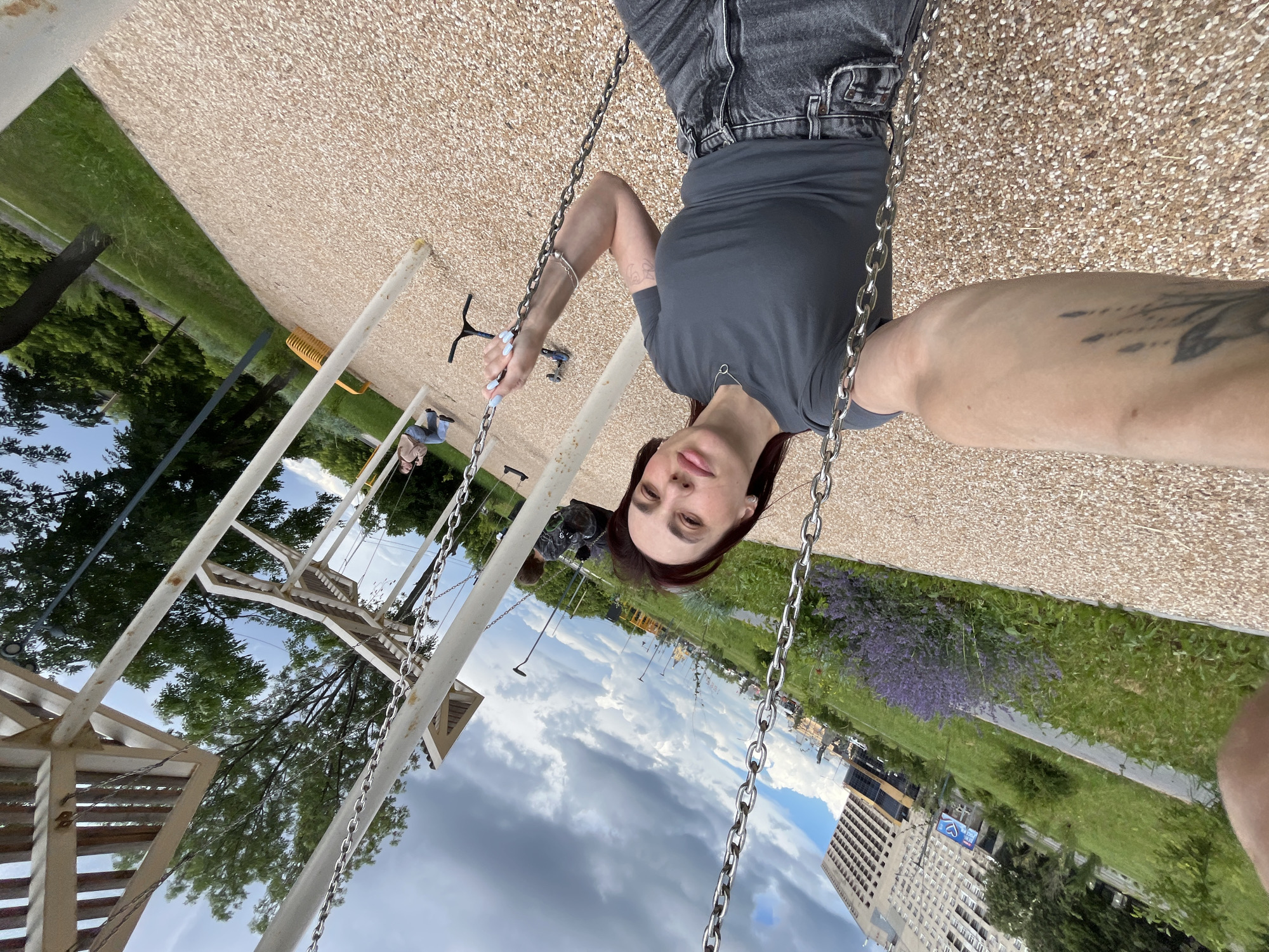 A woman is taking a selfie while sitting on a swing in a park. She has dark hair and is wearing a grey t-shirt and black jeans. She is wearing a silver necklace and a silver bracelet. Her left arm, which is visible in the foreground, has a tattoo. She is sitting on a swing with metal chains. In the background, there are other swings, trees, a large building, and a cloudy sky. The park has a ground cover of small, light-colored pebbles. A child is visible on another swing in the background. The location is a park in Nizhny Novgorod, Russia.