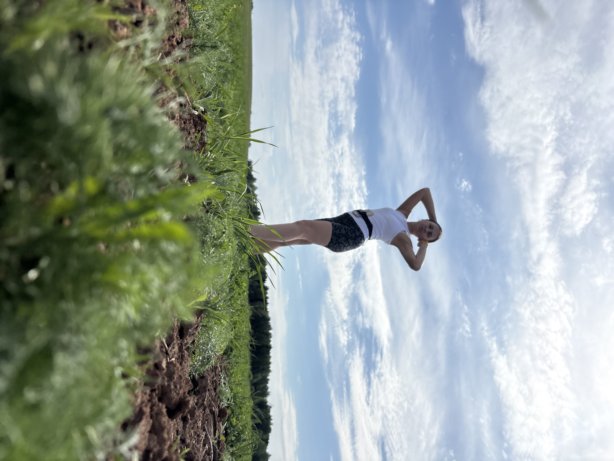 A young woman is standing in a field in Чумна, Russia. She is wearing a white tank top and patterned shorts. Her hands are clasped behind her head. The background is a blue sky with scattered white clouds. The field is covered in green vegetation. The ground appears to be a mix of dirt and grass.