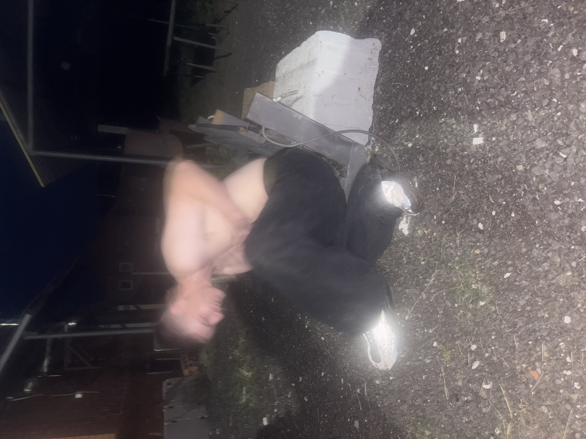 A male with light skin and short, light-colored hair is captured in a motion blur, appearing to be mid-movement while squatting. He is shirtless, wearing dark pants and white athletic shoes. His head is bowed, and his hands are near his abdomen. The background suggests an outdoor setting at night, with indistinct structures and dark foliage. To the right of the individual, a large, rectangular concrete block is visible, with a dark, metallic object leaning against it and a black cord attached. The ground is covered with gravel and scattered small stones.