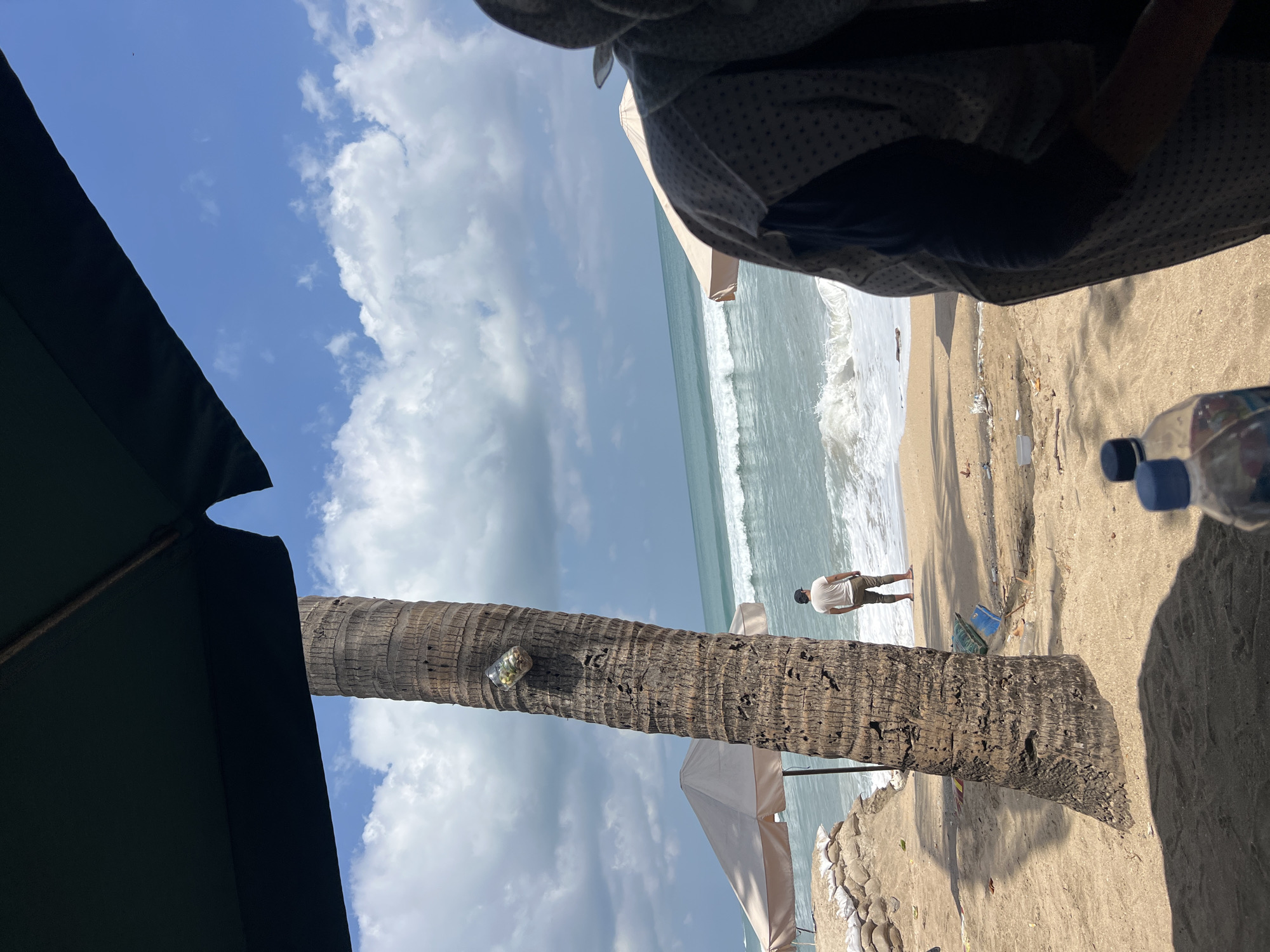 The image captures a beach scene in Kuta, Indonesia, on a day with scattered clouds and a bright sky. In the foreground, a large, textured palm tree trunk dominates the left side, with a glass bottle containing some contents attached to its side. To the right, the blurred upper body of a person, wearing a patterned hooded garment, is visible.
The middle ground of the image reveals a sandy beach with several sandbags and a closed umbrella in the background on the left, suggesting some form of coastal protection or setup. Further back, a person in a light-colored t-shirt and dark pants stands facing the ocean. The ocean itself is active with moderate waves breaking on the shore. Debris is scattered on the sand near the water's edge.
The background shows a calm sea meeting a sky filled with fluffy white clouds. The overall impression is a somewhat weathered, yet lively, beach environment.
