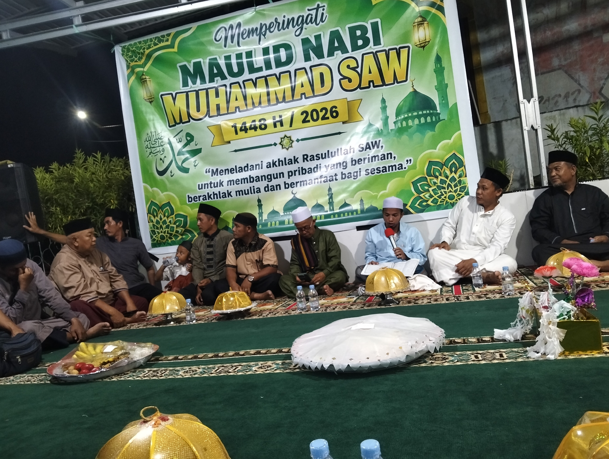 The image captures an indoor gathering, likely a religious event, at a location in Petapa, Indonesia. A large green banner with Arabic calligraphy and images of mosques is prominently displayed, indicating a commemoration of "Maulid Nabi Muhammad SAW 1448 H / 2026." This translates to the celebration of the Prophet Muhammad's birthday.
Several men are seated on a green carpet, some wearing traditional Islamic attire such as peci (caps) and thobes. They appear to be listening or participating in the event. In the foreground, there are several objects arranged on the carpet, including platters of food (likely fruit), decorative golden covers resembling lampshades, and bottles of water. One man in the background is speaking into a microphone, holding a book. The environment appears to be a temporary setup, possibly in a community hall or an outdoor covered area, suggested by the exposed ceiling structure and lighting. The overall atmosphere suggests a respectful and communal observance.