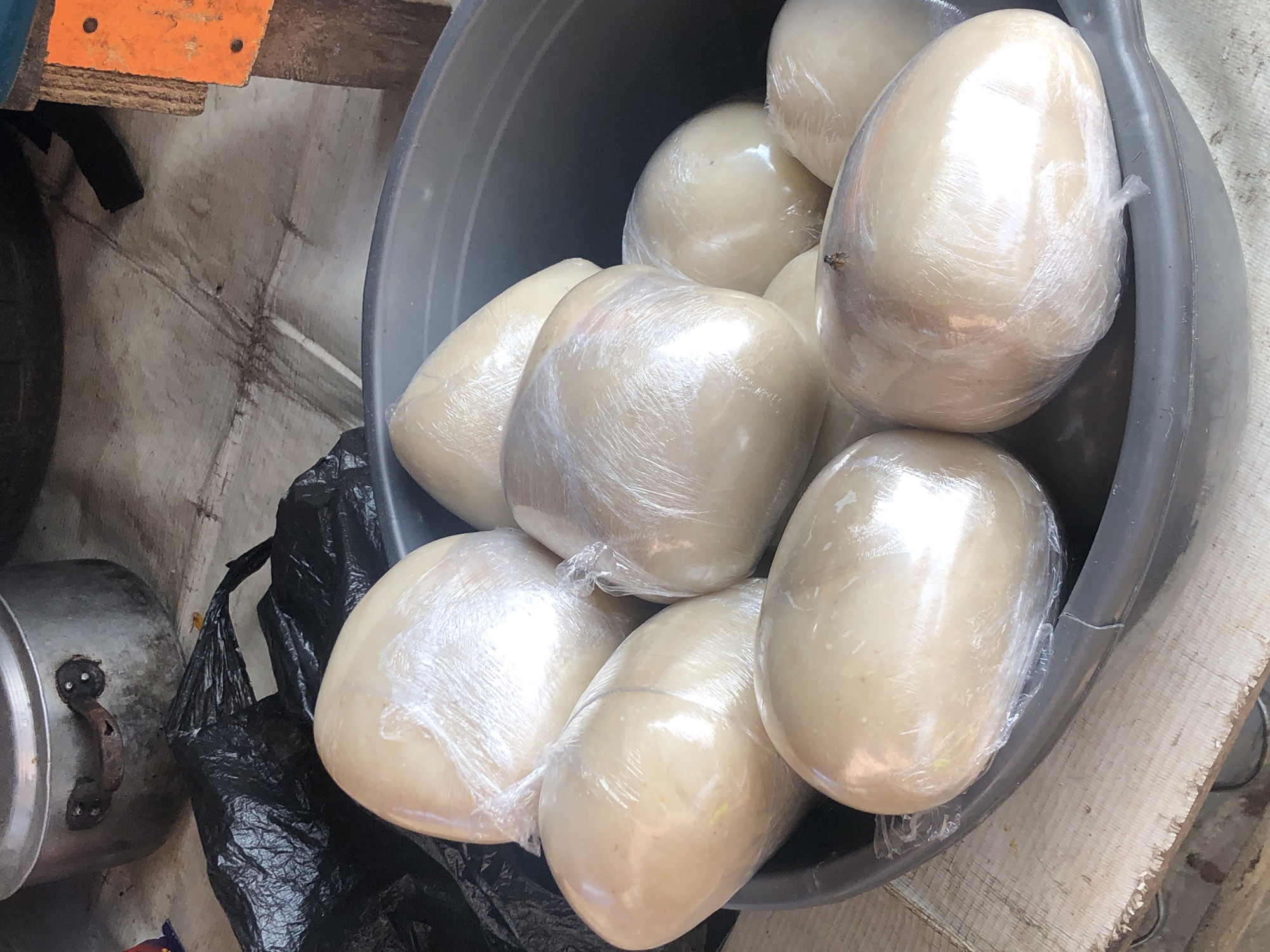 In Ikeja, Nigeria, a dark gray plastic tub holds several large, oval-shaped food items, possibly fufu or pounded yam, individually wrapped in clear plastic. The food is light beige in color and has a smooth, slightly shiny surface. The tub is filled with these wrapped portions, creating a piled effect. To the left of the tub, a crumpled black plastic bag is visible. In the background, a large, metallic cooking pot with two handles sits on a surface that appears to be made of concrete or stone. Part of an orange and wooden structure is also visible on the right side of the image. The overall scene suggests a food vendor's stall or a preparation area for a meal.