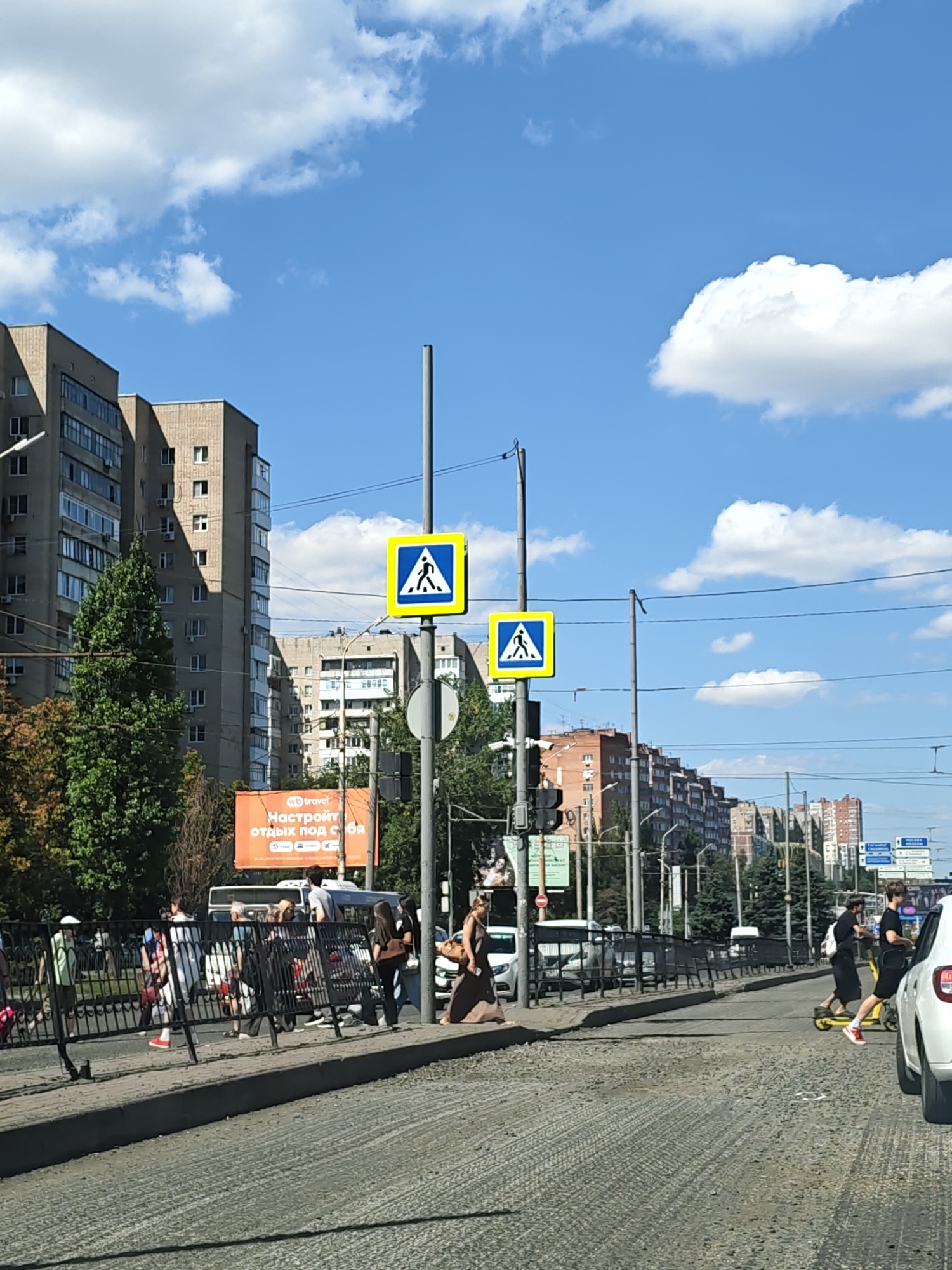 The image depicts a street scene in Rostov-on-Don, Russia, on a sunny day with scattered clouds. In the foreground, a road under construction is visible, with a textured, uneven surface. A white car is parked on the right side. Further down the street, a crosswalk is marked, with two pedestrian crossing signs mounted on poles. Several people are present: some are walking on the sidewalk or crossing the street, while others are near a black metal fence. One individual is riding a yellow electric scooter. In the background, there are multi-story apartment buildings and trees. An orange billboard with Russian text is also visible. Power lines stretch across the sky. The overall scene suggests an urban environment with ongoing infrastructure work.