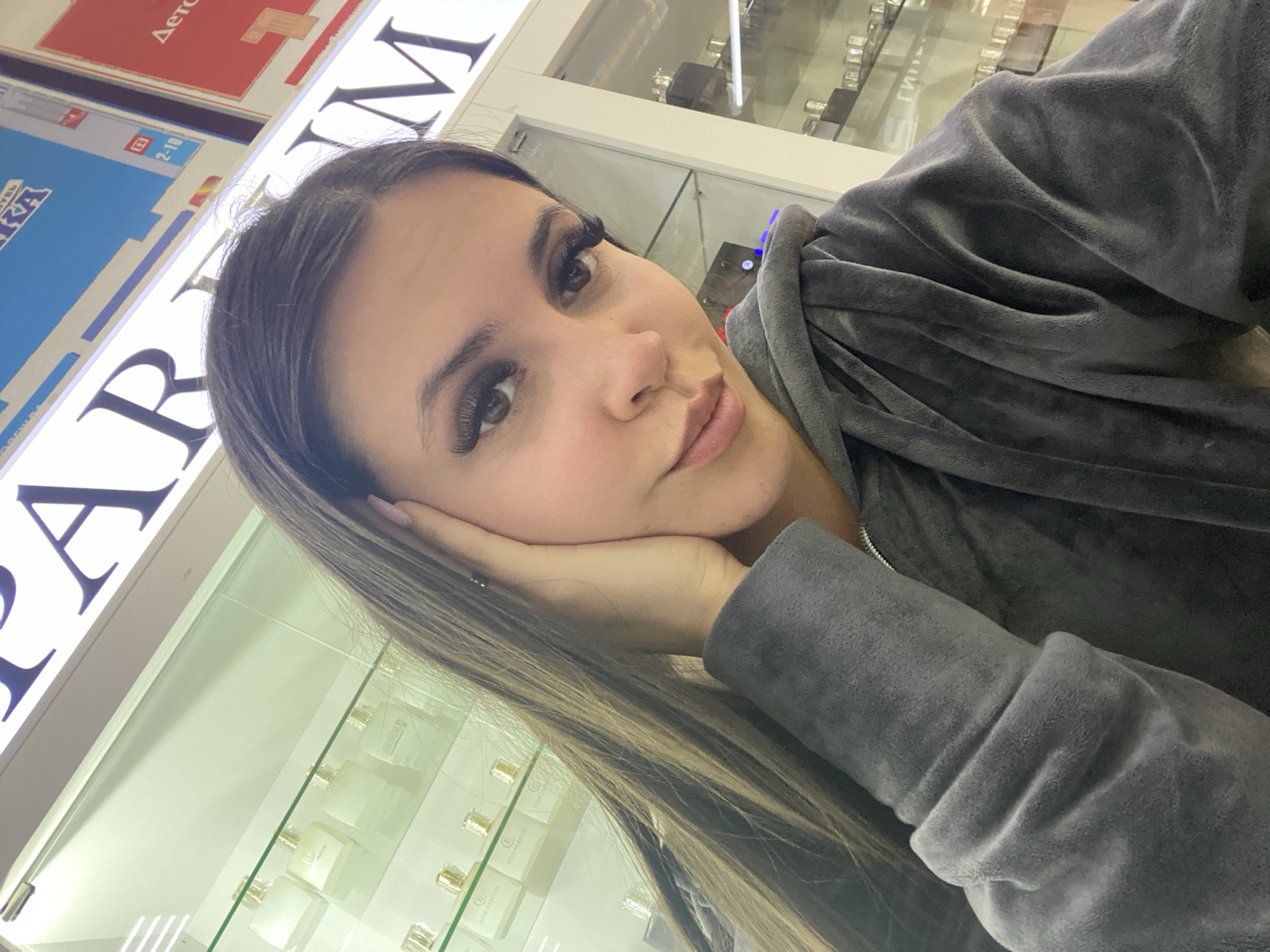 A young woman poses in a store that sells perfume, as indicated by the word "PARFUM" visible behind her. She wears a gray velour hooded sweatshirt and leans her face on her left hand. The scene is likely located in Chaikovsky, Russia, as it is tagged as such. Behind her is a glass display case containing small, elegant perfume bottles, suggesting a retail environment.