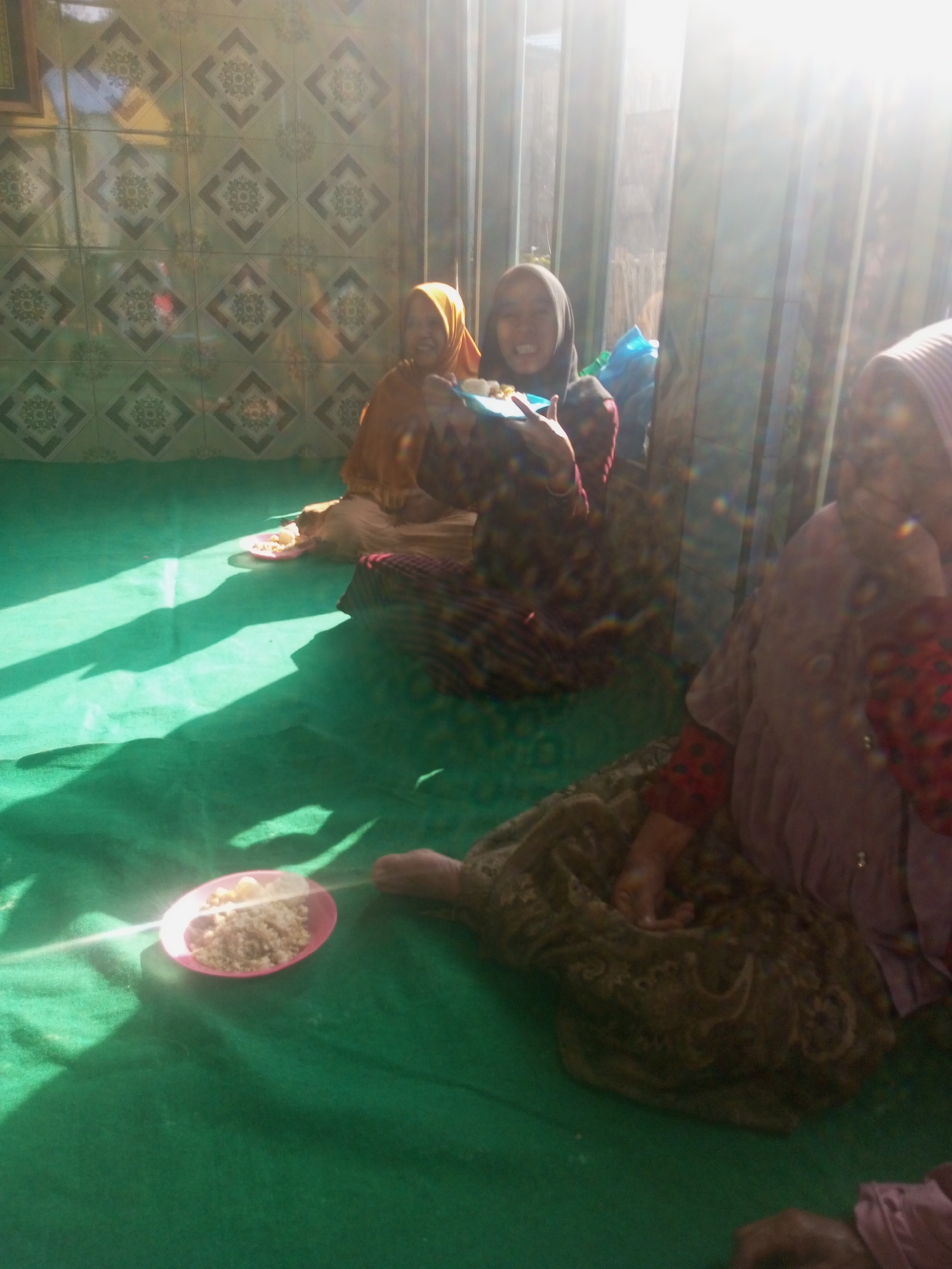 In a room in Andongsili, Indonesia, three women are visible, seated on a green carpeted floor. The room is illuminated by strong sunlight streaming in from the right side of the frame. The back wall is covered in patterned tiles.
The woman on the far left is wearing a yellow hijab and a brown outer garment. She is smiling and appears to be eating from a small pink plate in front of her, which contains what looks like rice.
To her right is another woman wearing a dark hijab and a colorful patterned garment. She is also smiling, and her hands are visible. She is holding a blue object and looking towards the left.
On the far right, the third woman is partially visible, wearing a light purple head covering and a patterned red and dark garment. Her hands are also visible, resting on a patterned fabric. The scene suggests a casual gathering or meal.