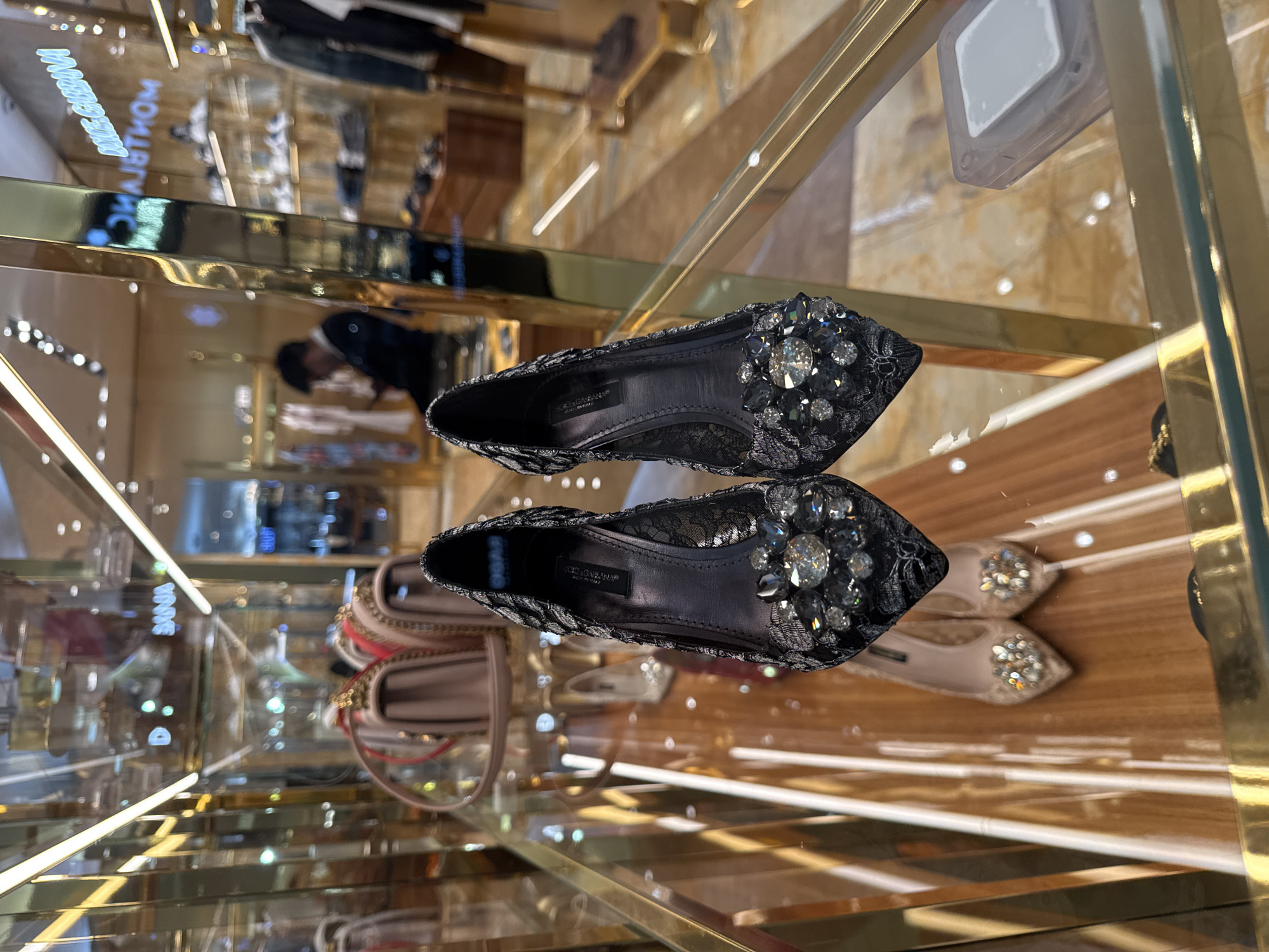 The image shows two pairs of Dolce & Gabbana shoes displayed on a glass shelf with a gold-colored metal frame, typical of a retail store. The foreground pair of shoes are black lace flats with jewel embellishments. Partially visible is another pair, likely a similar design but in a lighter color. The reflective nature of the glass shelf creates multiple reflections of the shoes and other items in the store, including other clothing racks and a handbag. The store location is Puebla, Mexico, as determined from storefront signage.