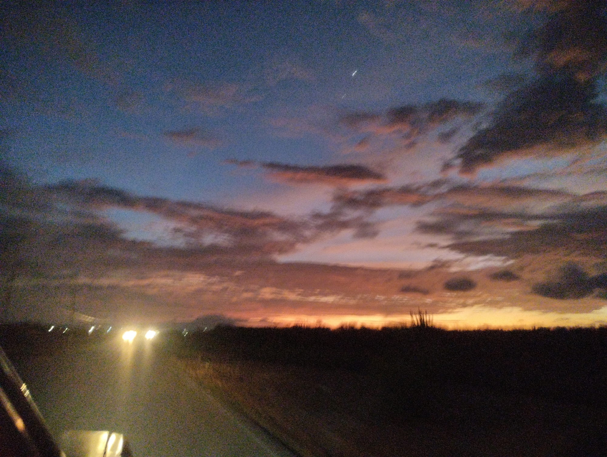 The image depicts a twilight scene from within a vehicle moving along a road in La Guaira, Venezuela. The sky is a gradient of deep blue to a soft orange, with scattered clouds tinged with pink and yellow hues. The horizon is marked by a dark silhouette of land, likely vegetation or low hills. In the distance, on the road, the headlights of other vehicles create bright, elongated streaks of light. The foreground shows a portion of the vehicle's interior, including part of the windshield and dashboard. The overall impression is one of a journey at dusk, with the natural beauty of the sky contrasting with the artificial lights of passing traffic.