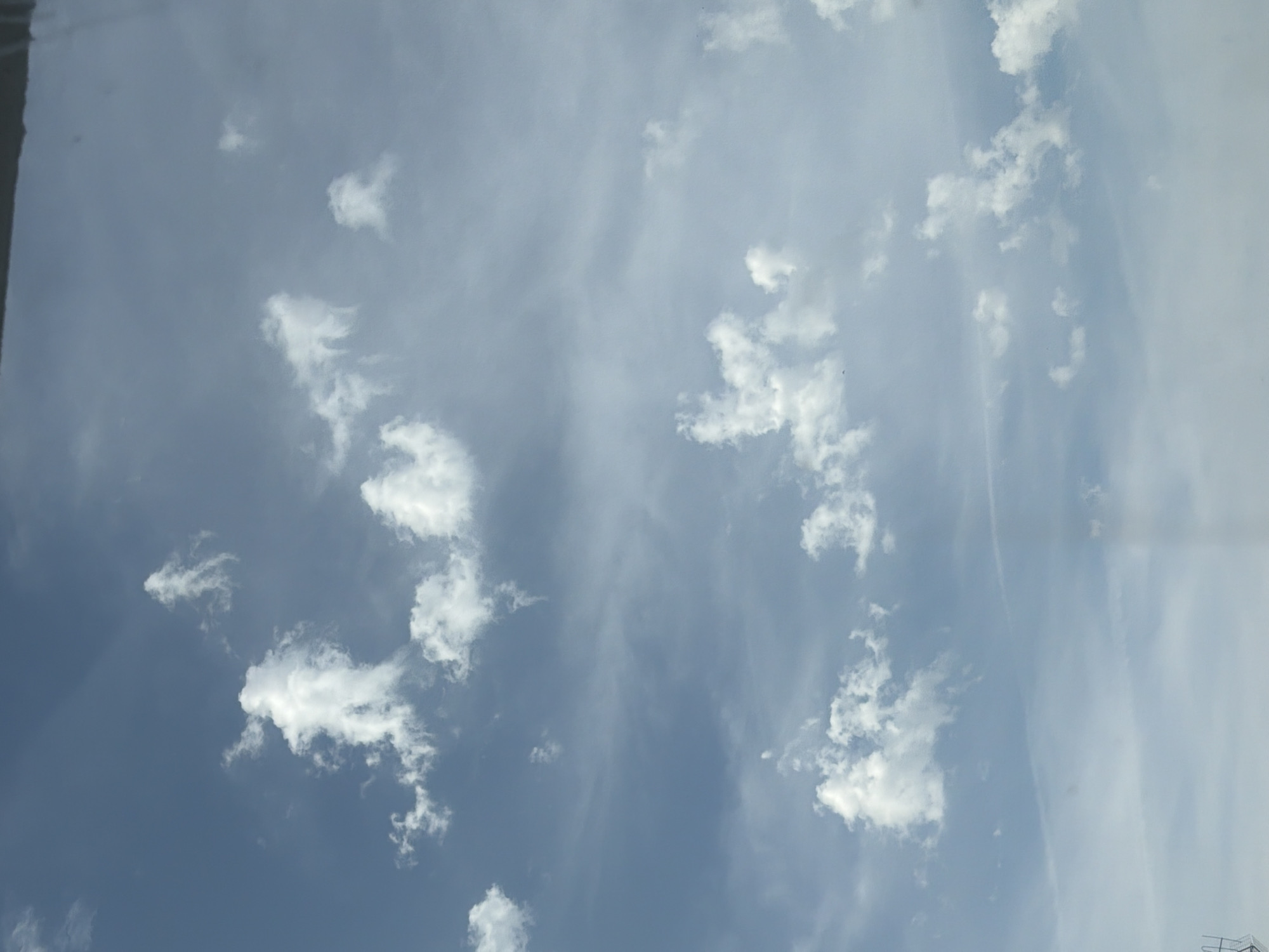 The image is a vertical, full shot of a pale blue sky with scattered white cumulus clouds. The clouds are irregularly shaped and appear to be drifting across the sky. Some clouds are wispy and thin, while others are more dense and fluffy. The overall impression is of a bright, possibly sunny day, but with cloud cover. There are no people, subjects, or objects clearly visible in the sky. The bottom edge of the image shows a slight obstruction, possibly a window frame or a building edge, which is out of focus. No specific location within the city of Burgas, Bulgaria, can be identified from this image alone.