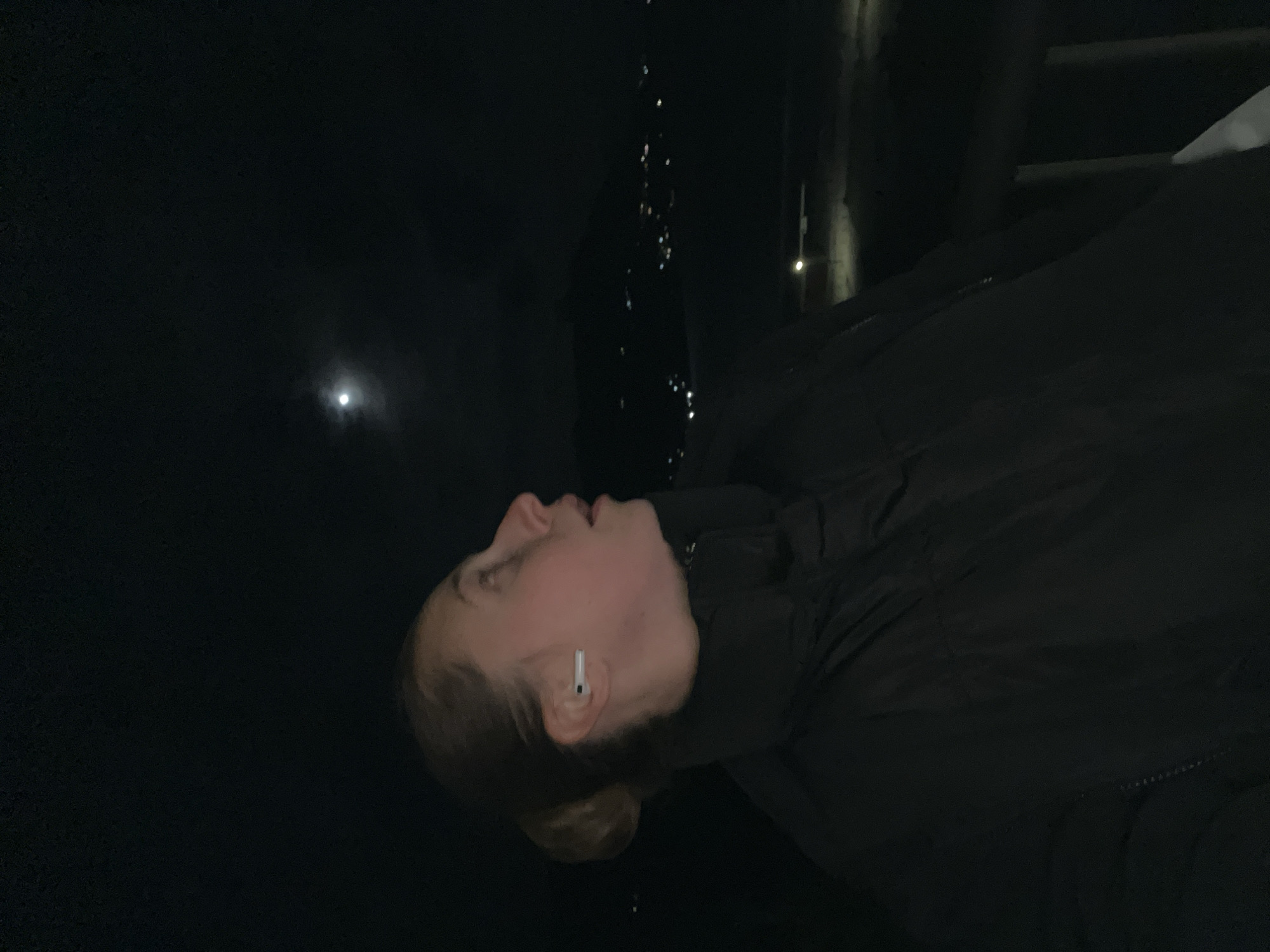 A low-light photograph shows a person wearing a dark jacket and an earbud, looking up at the moon. The individual's head is tilted back and to the left. In the background, there are faint city lights, and a partial structure can be observed. Based on the prompt, the location is inferred to be the city of Boreti, Montenegro.