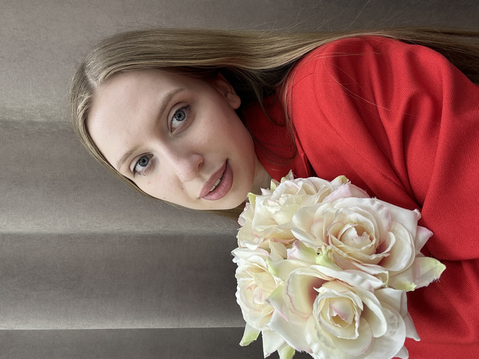 This image, located in Chaikovsky, Russia, presents a close-up portrait of a young adult female. She possesses fair skin, blue eyes, and long, straight light-brown hair parted slightly to the left. Her head is tilted to her right, and she is looking directly at the camera with a subtle, closed-lip smile. She is wearing a bright red blazer or jacket, visible across her shoulders and upper torso. Held in her left arm, resting against her chest and left shoulder, is a compact bouquet of approximately eight to ten light-colored artificial roses. These flowers are primarily off-white to pale pink, with hints of light green at the base of some petals. The background is a uniform, vertically textured, dark beige or light grey fabric, resembling a curtain or upholstered wall. No other individuals or distinguishing objects are visible.
