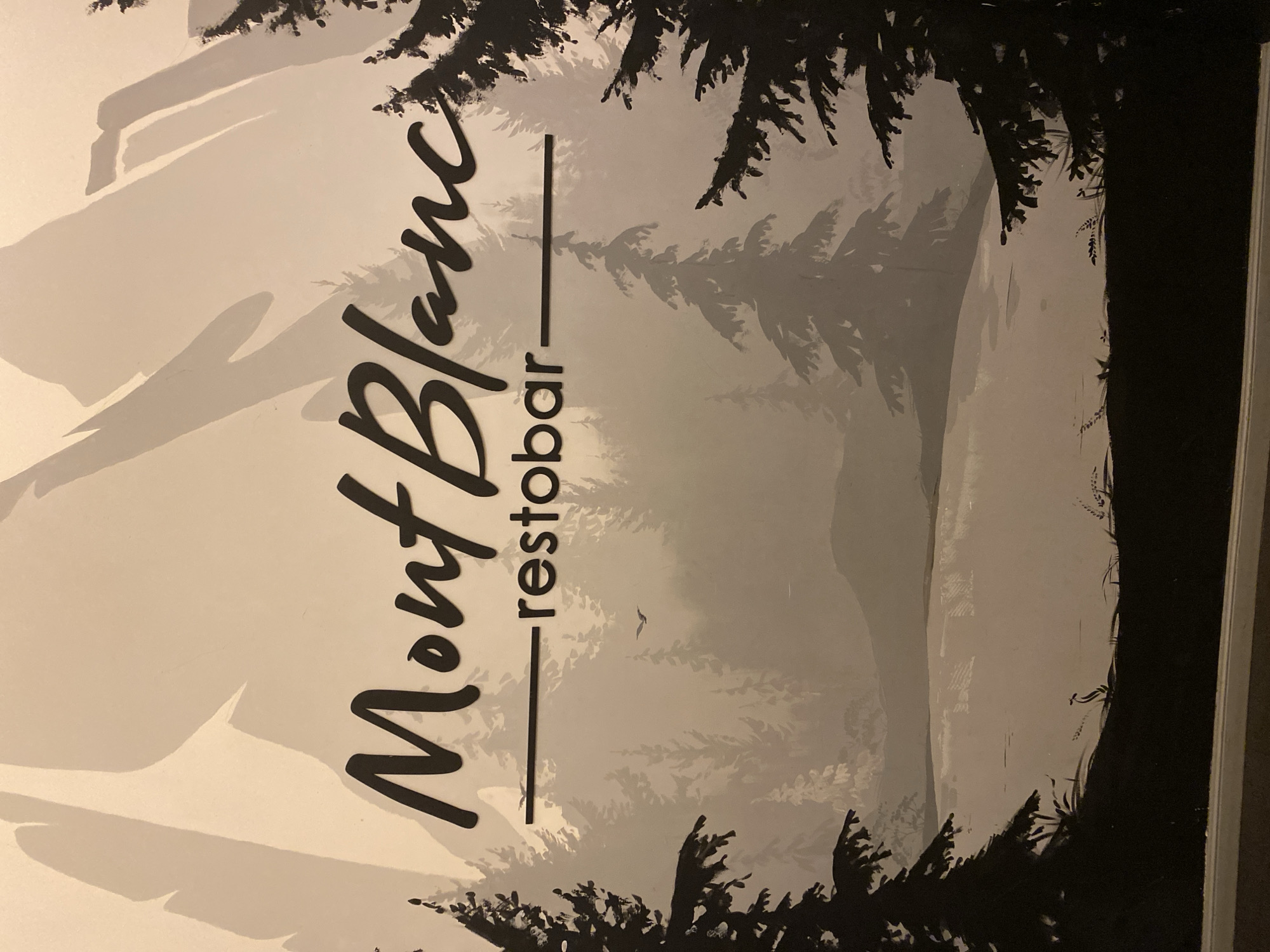 The image shows a stylized mural depicting a mountain landscape with trees and a body of water. The text "Mont Blanc - restobar" is prominently displayed in a script font. The mural is monochromatic, primarily in shades of grey and black, with a pale background representing mountains. There are silhouetted pine trees in the foreground and middle ground, and a serene lake or river in the lower portion of the image. A small bird is visible in flight in the distance. The overall aesthetic is artistic and suggestive of a serene, possibly upscale, establishment. There is no indication of the specific location within Petropavl, Kazakhstan, in the image itself.
