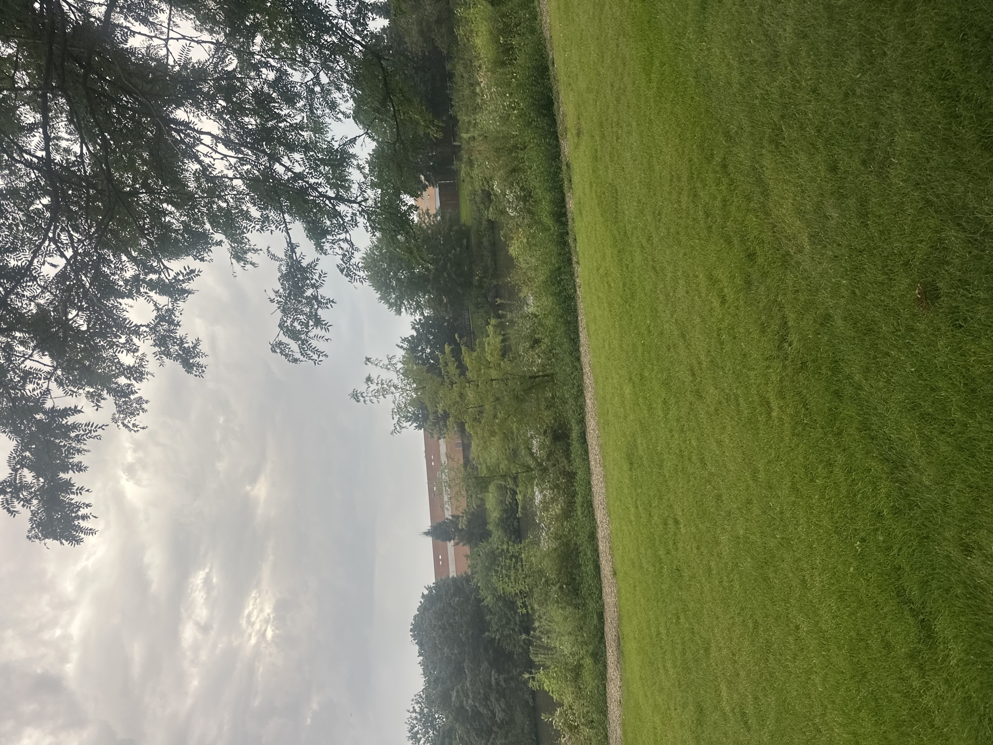 The image depicts a serene natural landscape with a large building in the background. In the foreground, a lush green lawn stretches across the frame, suggesting a park or recreational area. A gravel path borders the lawn and leads towards a body of water. Beyond the path, a dense collection of trees and vegetation lines the water's edge. A portion of a building, characterized by a reddish-brown facade and a dark roof, is visible above the trees. The sky is overcast with soft, diffused light. The scene appears to be a tranquil outdoor setting, possibly part of a park or green space within a suburban environment. The specific location of Schaumburg, United States of America, is not discernible from the visual elements present in the image.
