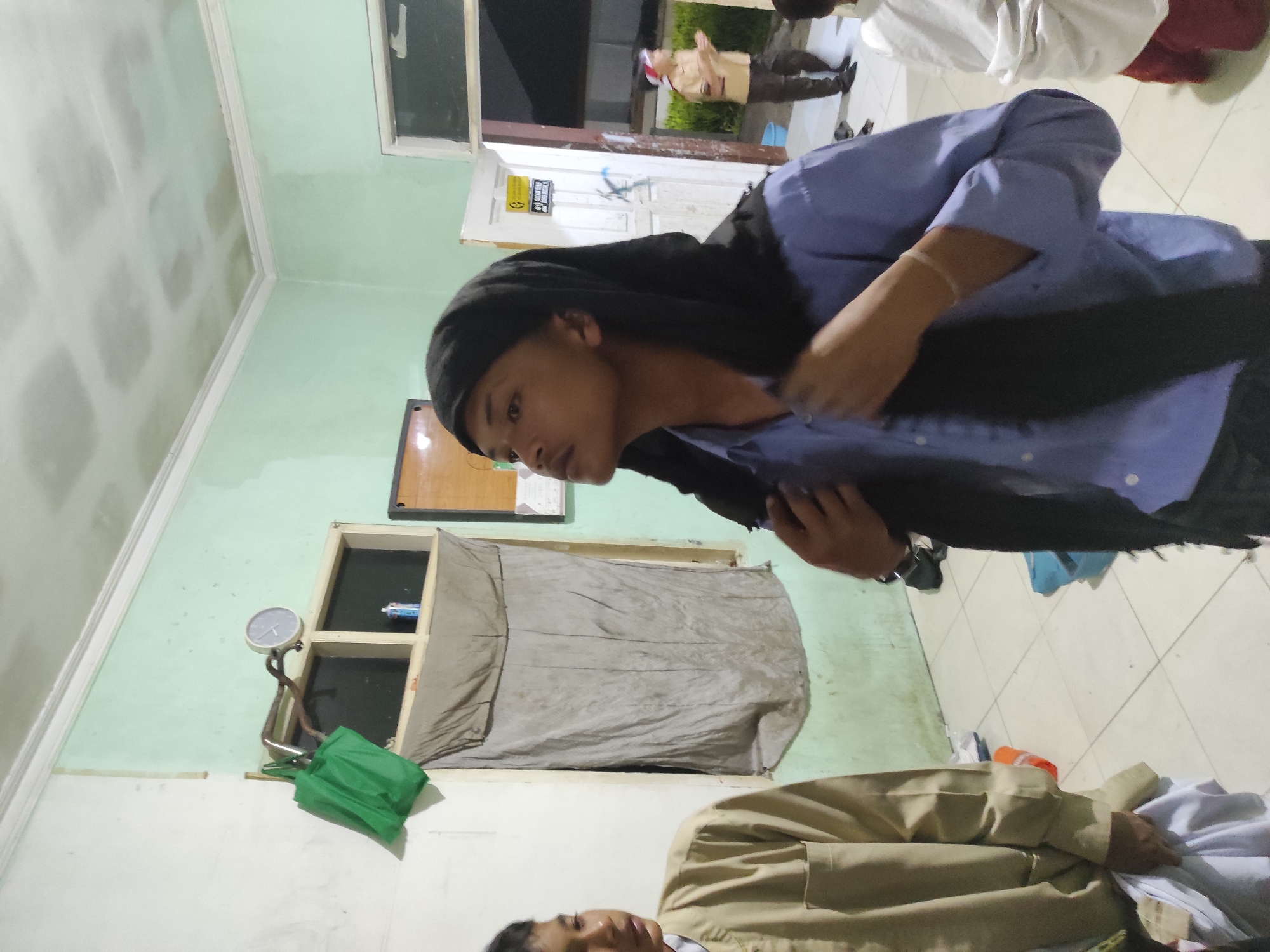 The image depicts an indoor scene, possibly a room in Sumedang, Indonesia. In the foreground, a young person wearing a blue shirt and a black head covering is adjusting their clothing. To their left, another young person in a tan shirt stands partially visible, holding a white cloth. The room has pale green walls, a white ceiling with visible ceiling panels, and a tiled floor. Various objects are present, including a green bag hanging on a rack near a window with a gray curtain. A clock is mounted on the wall above the window. In the background, an open doorway reveals another person in a tan uniform with a red and white head covering, and the suggestion of an outdoor area beyond. The lighting is dim, creating shadows and contributing to a subdued atmosphere. The overall impression is of a casual gathering or activity within a utilitarian space.