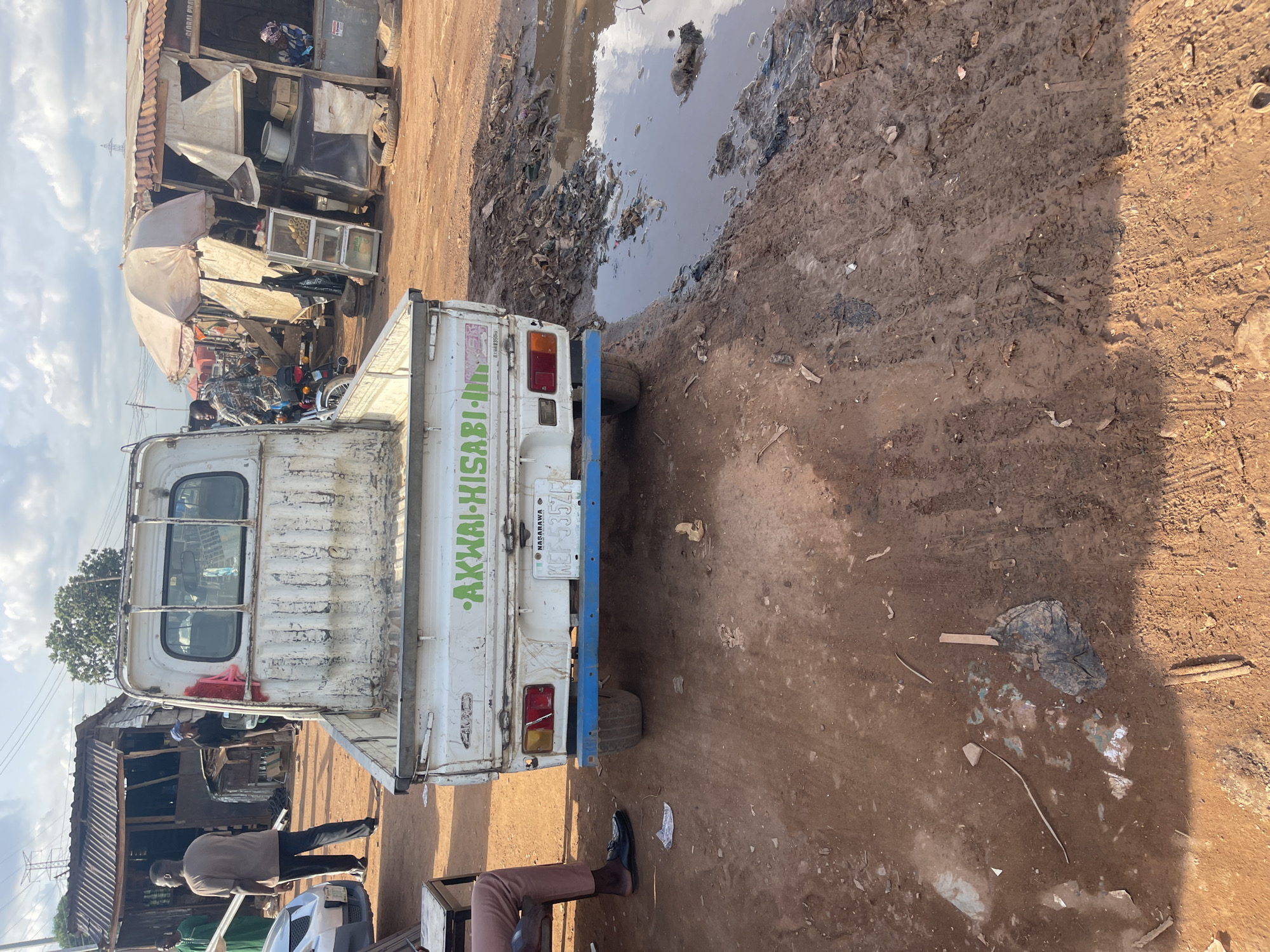 The image captures a scene in an area with muddy terrain, likely in or near Abuja, Nigeria, as indicated by the NASARAWA state license plate "KEF-535ZF" on the back of a small, white truck. The truck, bearing the words "AKWAI-HISABI-IN...", appears to be in a state of disrepair. Next to the truck, there is a water puddle containing some trash. Various structures or stalls are visible in the background. People are seen both near these structures and near the vehicle. The overall environment has an unkempt and somewhat impoverished look.