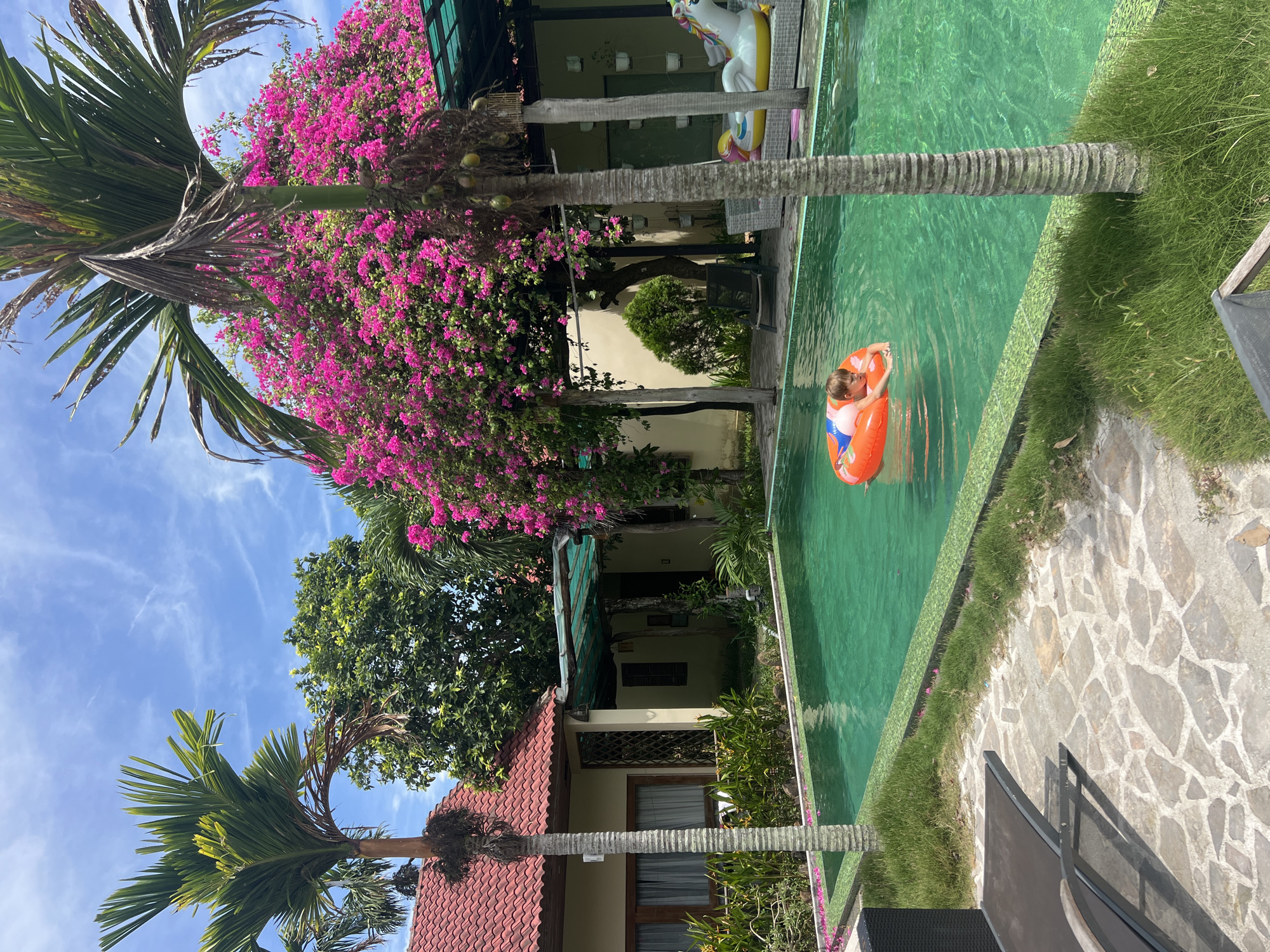 A young child is swimming in a turquoise pool, wearing an orange inflatable ring. The child is looking down at the water. The pool is surrounded by lush green grass and a stone pathway. In the background, there are palm trees, a building with a red tiled roof, and vibrant pink bougainvillea flowers. An inflatable unicorn float is visible near the edge of the pool. The scene appears to be a resort or private residence in Hoi An, Vietnam, indicated by the architectural style and tropical vegetation. The sky is a clear blue with some wispy white clouds.