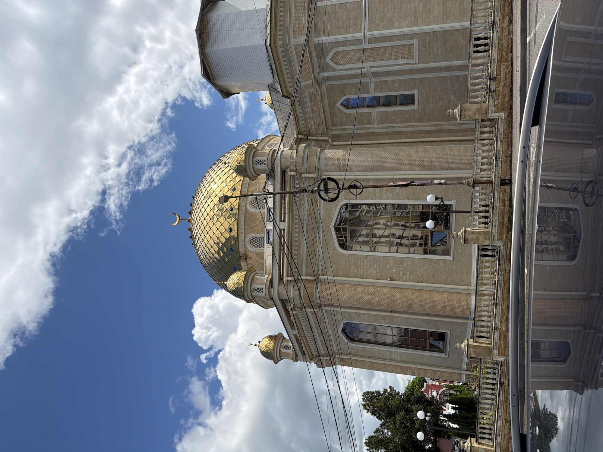 The image captures the exterior of the Sunna Mosque in Kislovodsk, Russia, under a bright blue sky with scattered clouds. The mosque features a prominent golden dome, adorned with a crescent moon symbol at its apex. Smaller golden domes and minarets also punctuate the architecture. The building's facade is constructed from light-colored stone with decorative brickwork. A white stone balustrade runs along the foreground, separating the mosque from a street. Electrical wires crisscross the scene in front of the mosque. In the lower portion of the image, the reflection of the mosque and the sky is visible on the dark, glossy surface of a car roof. Trees and streetlights are also present to the left of the mosque. The overall scene depicts a religious building in an urban setting on a clear day.