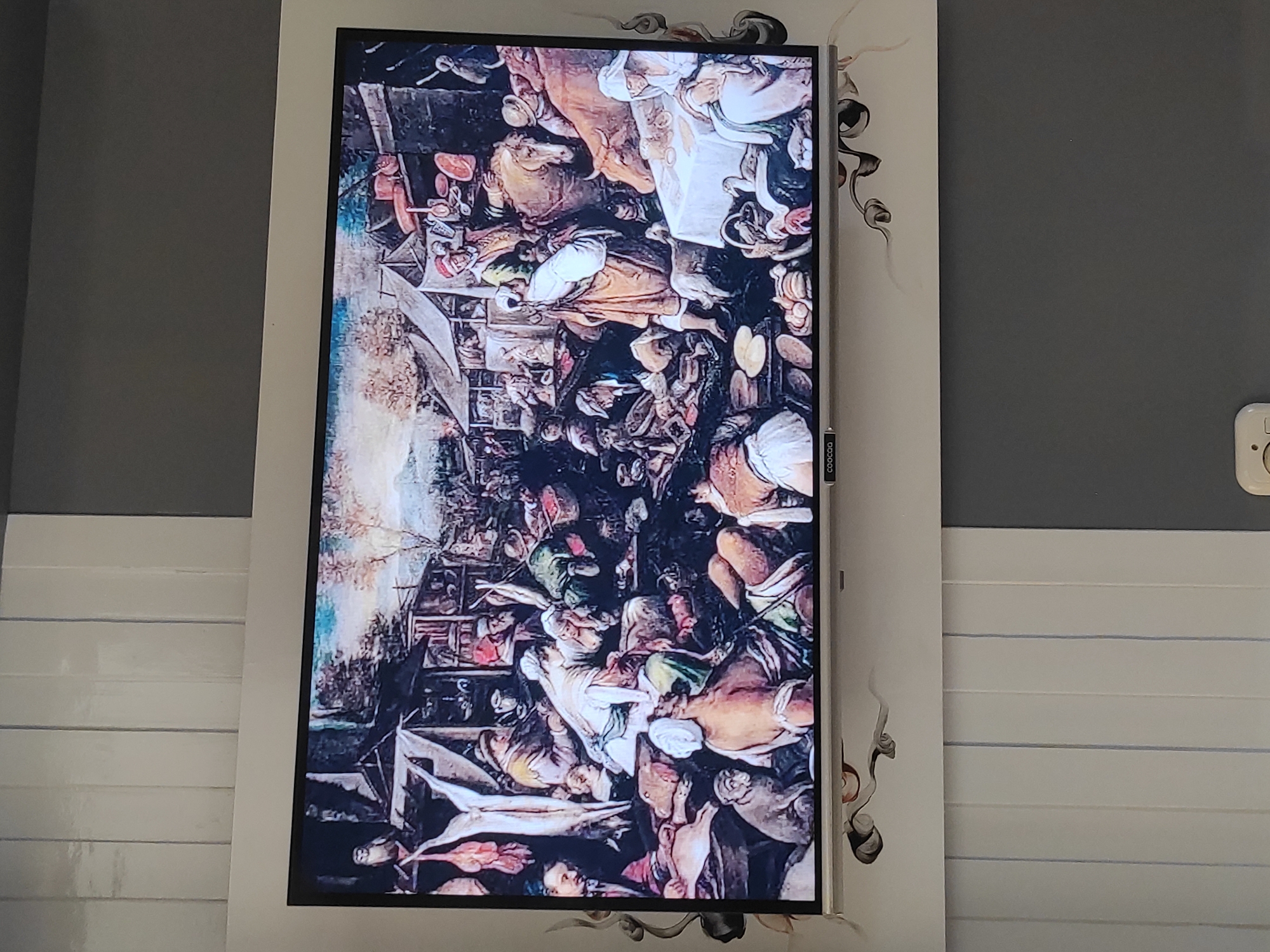 The image displays a painting on a television screen, depicting a crowded marketplace scene, possibly from a historical period. The painting features numerous figures engaged in various activities, including selling goods, interacting with each other, and tending to animals. There are stalls laden with food items, including meats and produce. The overall atmosphere appears lively and bustling. The television is mounted on a wall with vertical paneling, and the surrounding environment suggests an indoor setting. The logo "COOCAA" is visible on the bottom bezel of the television. The painting itself exhibits a detailed and realistic style. The question about the location in Sumedang, Indonesia, cannot be answered from the visual information provided in the image.