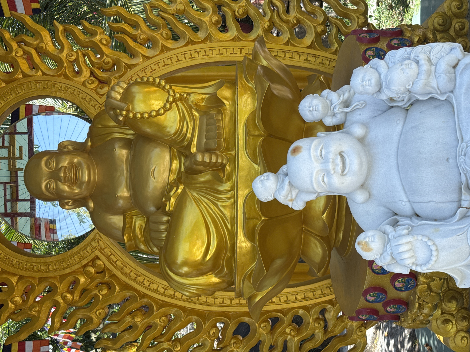 The image captures two statues of Buddha, likely photographed at a temple or shrine in Bac Nha Trang Ward, Vietnam. The statue on the left is golden, with intricate detailing in the ornate design around its circumference. A mirror-like surface partially reflects the surrounding environment. The right-hand statue is white, depicting a Buddha figure with smaller figures climbing on it. The ground or base of the statue displays small, round decorations with painted designs. The background shows green foliage, possibly from trees or bushes, suggesting an outdoor setting.