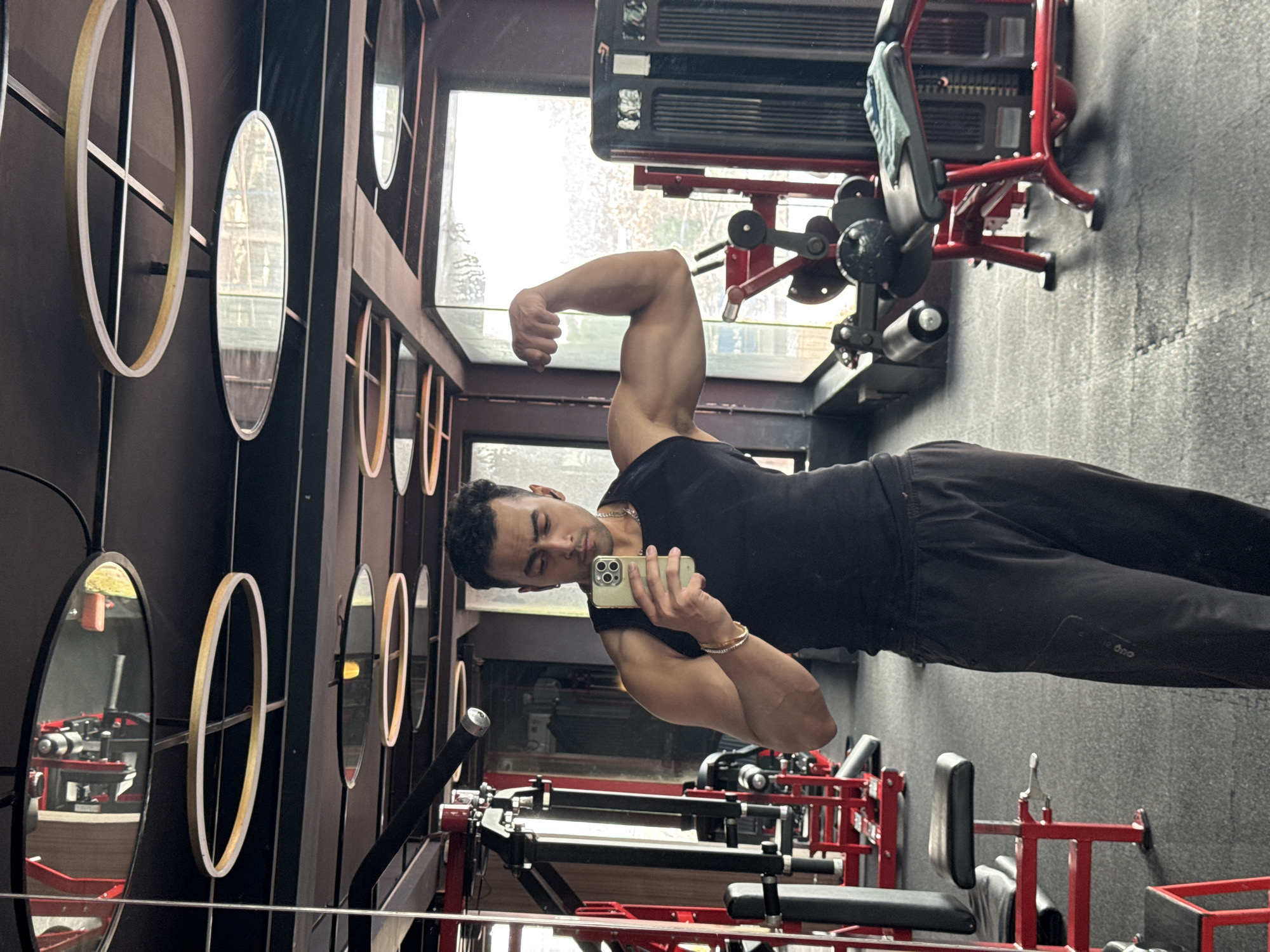 The image shows a young man taking a mirror selfie in a gym. He is wearing a black sleeveless shirt and black pants, flexing his bicep. The gym appears to be modern with red and black equipment, including weight machines and benches. The ceiling has circular light fixtures and mirrors. The overall impression is of a well-equipped, contemporary fitness center. This image is not located in Kolkata, India.