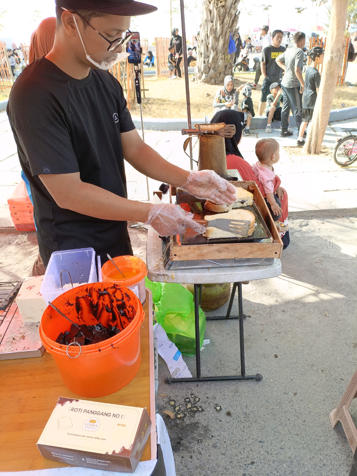 A street vendor wearing a black t-shirt, black cap, and a face shield is preparing grilled bread with a spatula and tongs. He is standing behind a food cart in what appears to be a public outdoor area. In front of him, on a table, are an orange bucket filled with a dark spread, a clear plastic container, a white block, and a box of "Roti Panggang No. 1" from "Lunrea Bakehouse". In the background, several people, including women and children, are visible. A young child sits on a woman's lap, observing the vendor. The environment includes a paved ground, some greenery, a palm tree, and other people in the distance, suggesting a busy public space, possibly a park or a street market. The scene is set in Bulukumba, Indonesia, with the context of a local food vendor operation.