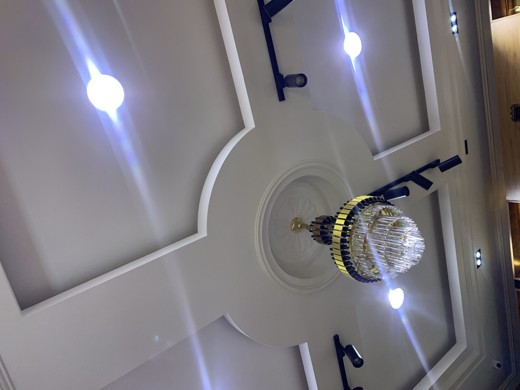 The image shows a modern ceiling with recessed lighting and a chandelier. The ceiling has multiple levels and decorative molding. Several recessed spotlights are visible, along with a track lighting system. A large, multi-tiered chandelier with a gold and crystal design hangs from the center of the ceiling. The lighting creates glare and lens flares throughout the image, obscuring some details. The location is Gura Top, Nigeria.