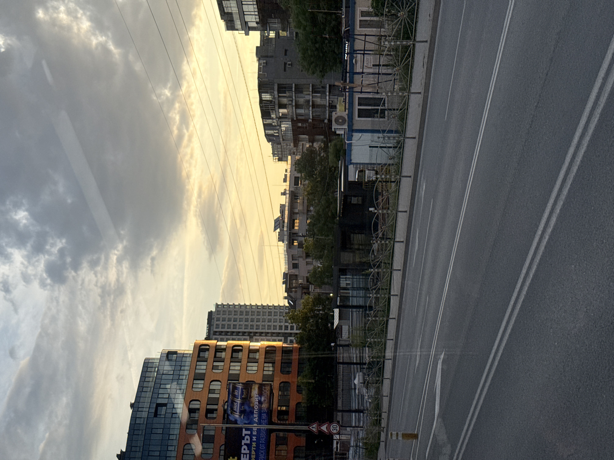 The image captures an urban scene in Sofia, Bulgaria, featuring a street view with a mix of modern and older buildings lining the road. On the left, a modern building complex with blue and orange facades catches the eye. A billboard with a local advertisement in Bulgarian is visible on one of the buildings. A tall, generic high-rise building stands in the background. A street with multiple lanes stretches towards the horizon, marked by clear white lane dividers. On the right side of the road, a decorative fence separates the sidewalk from the street. Power lines stretch across the sky, which is partly cloudy with a warm, golden hue from the sun. The overall perspective suggests the photo was taken from a moving vehicle or a high vantage point.