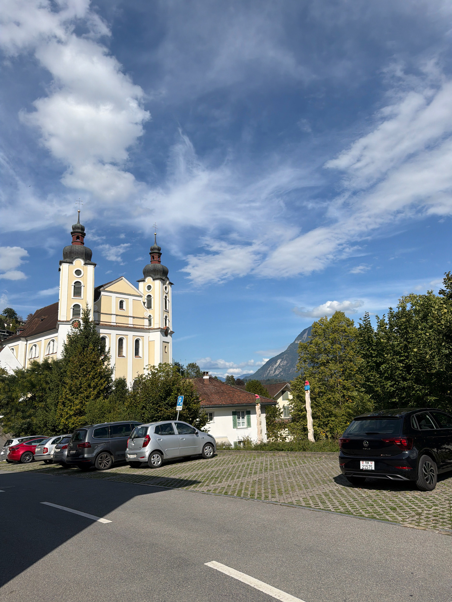 The image shows a church with two bell towers, painted yellow and white, against a blue sky with white clouds. In front of the church, there is a parking lot with several cars, including a black Volkswagen Polo, a silver hatchback, and a dark grey SUV. Trees and houses are visible in the background, as well as a mountain. The parking lot is paved with interlocking concrete blocks and grass. A blue parking sign is visible. The scene appears to be in a town or village, possibly in a mountainous region, on a sunny day.