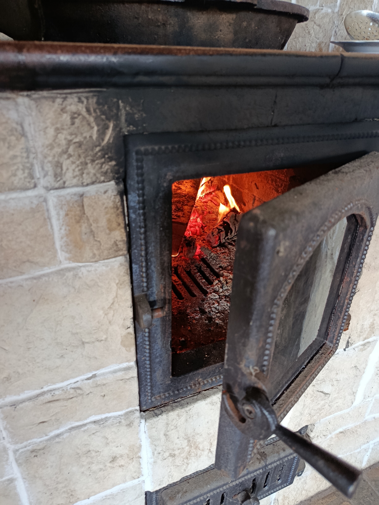 A close-up, eye-level shot captures a rustic, lit wood-burning stove set against a cream-colored brick wall. The stove's cast-iron door is ajar, revealing a vibrant fire within. Embers glow a fiery orange, with flames licking at charred wood. The door, adorned with a decorative beaded edge, hangs open, revealing a metal handle extending from its hinge. A dark metal grate is visible at the bottom of the stove, beneath the burning logs. Above the stove, partially visible, is a dark pot and a metal ladle resting on a white surface. The scene is likely in a kitchen or a similar domestic setting, possibly in the village of Misaylovo, Russia, given the context. The overall impression is one of warmth and traditional cooking.