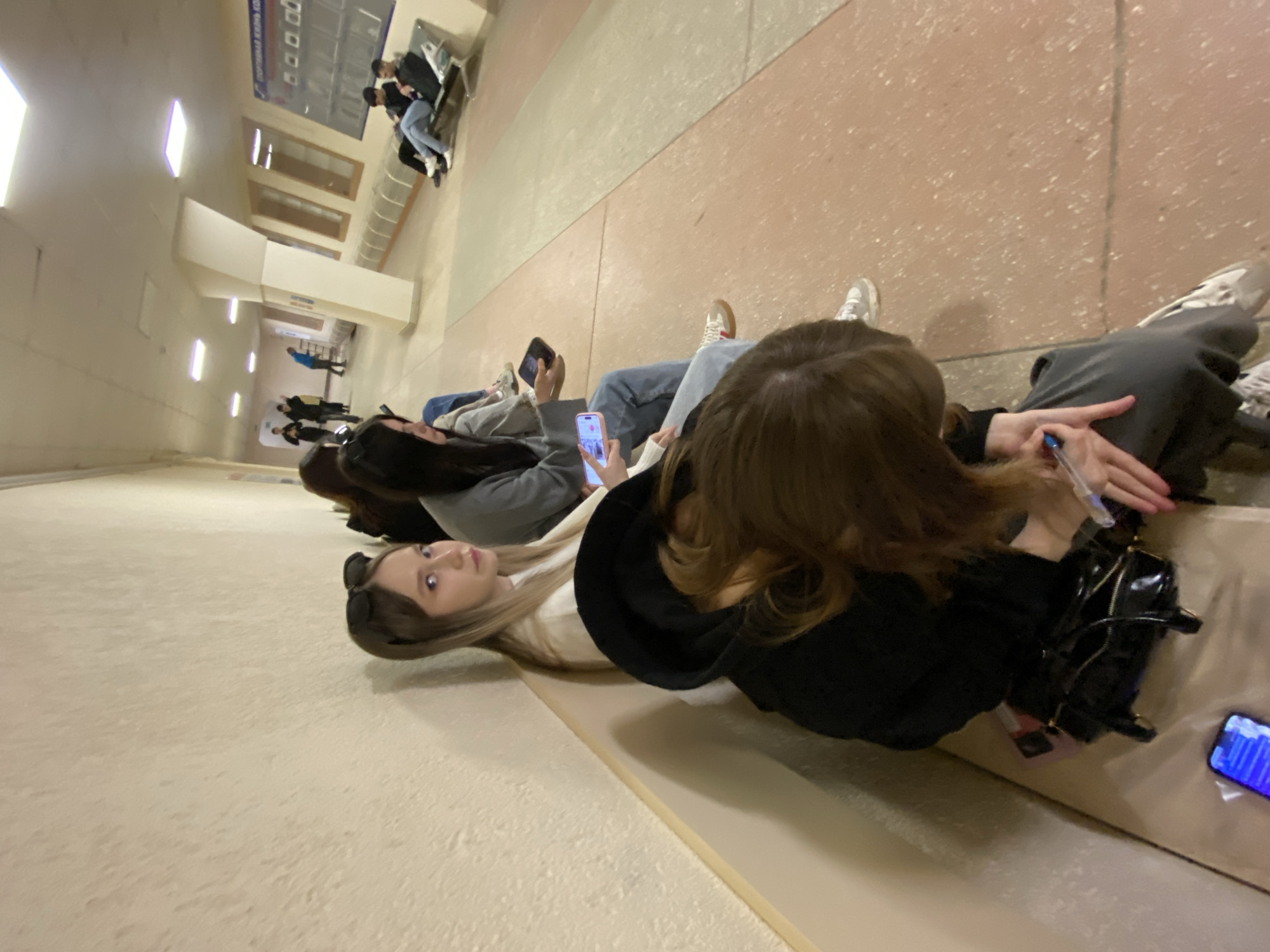 Several individuals are seen resting on a bench within a building. Many are using smartphones. The environment appears to be a waiting area or lobby, possibly at a transit center. The location may be in the city of Chaikovsky, Russia, given information visible on a sign in the image background.