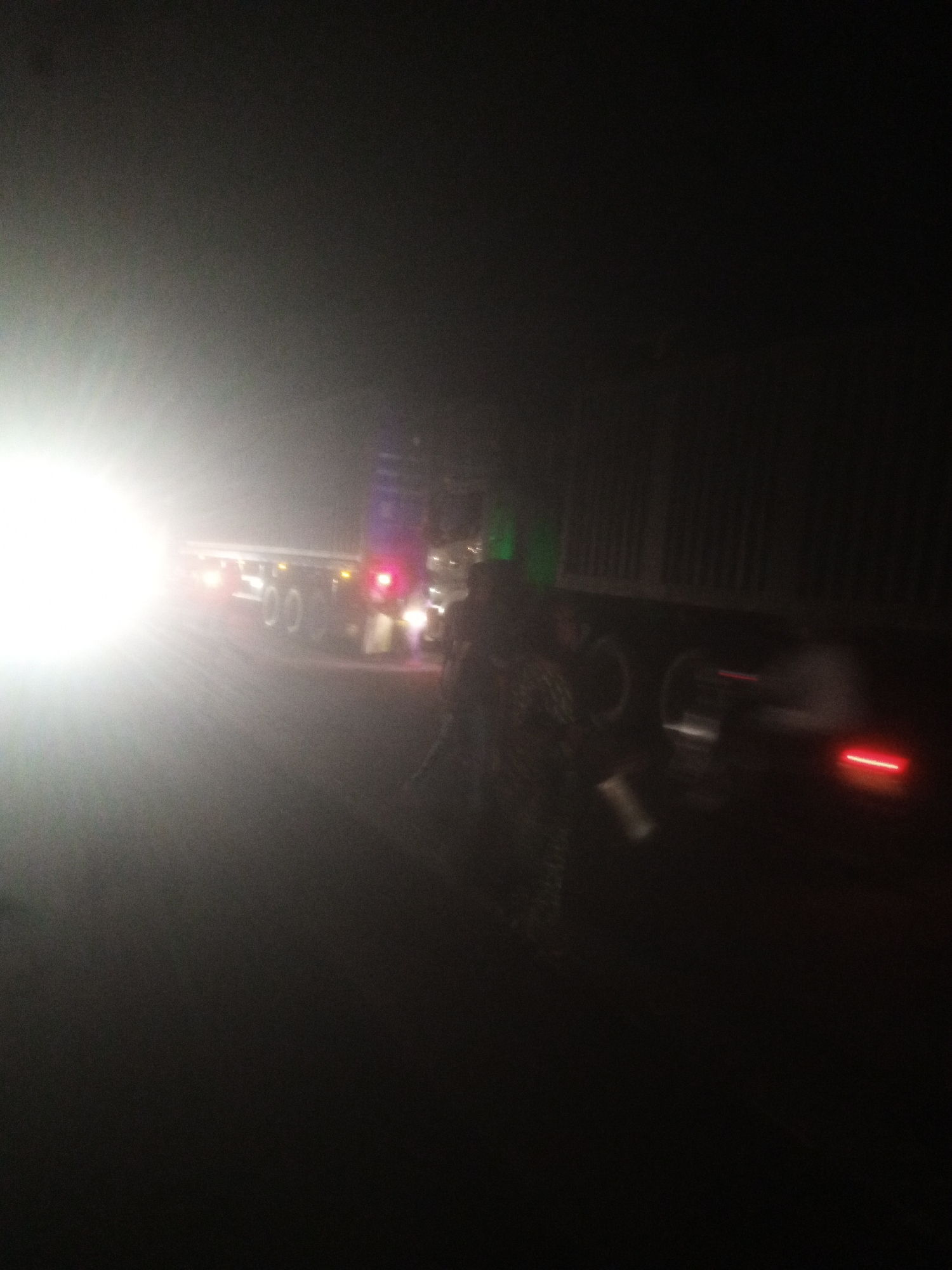 The image captures a nighttime scene on a dark road, likely in Nassarawa, Nigeria, where a large truck is parked. The truck's headlights are on, casting a bright, diffused light that dominates the left side of the frame. Red taillights are visible on the rear of the truck, indicating its presence on the road. The scene appears somewhat blurry, suggesting motion or low light conditions. A figure, possibly a person, can be seen near the truck, partially obscured by the glare of the headlights and the darkness. The overall environment is dark, with the truck and its lights being the most prominent features. The road surface is not clearly discernible due to the darkness and the lighting conditions.